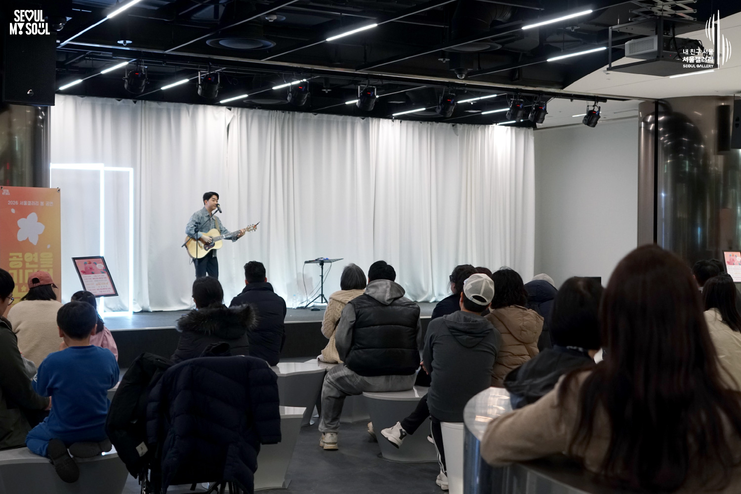 서울갤러리 '2026 서울갤러리 봄 공연' 무대에서 남성 싱어송라이터가 어쿠스틱 기타를 연주하며 노래하고 있고, 객석에 앉은 관객들이 공연을 관람하고 있는 모습.