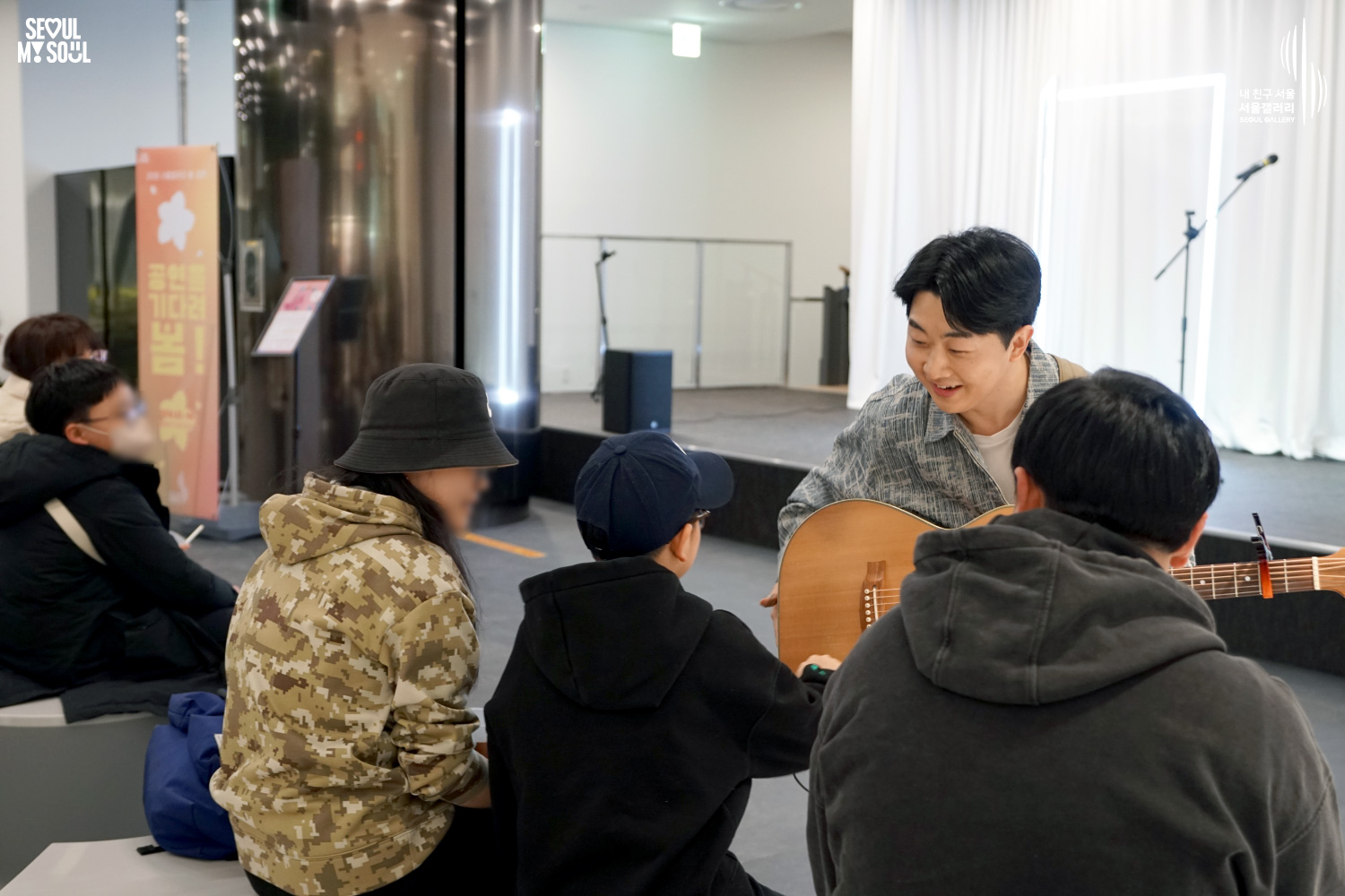 서울갤러리 '공연을 기다려 봄!' 행사에서 어쿠스틱 기타를 든 공연자가 무대 앞으로 내려와 관객들과 가까이서 미소 지으며 소통하고 있는 모습.