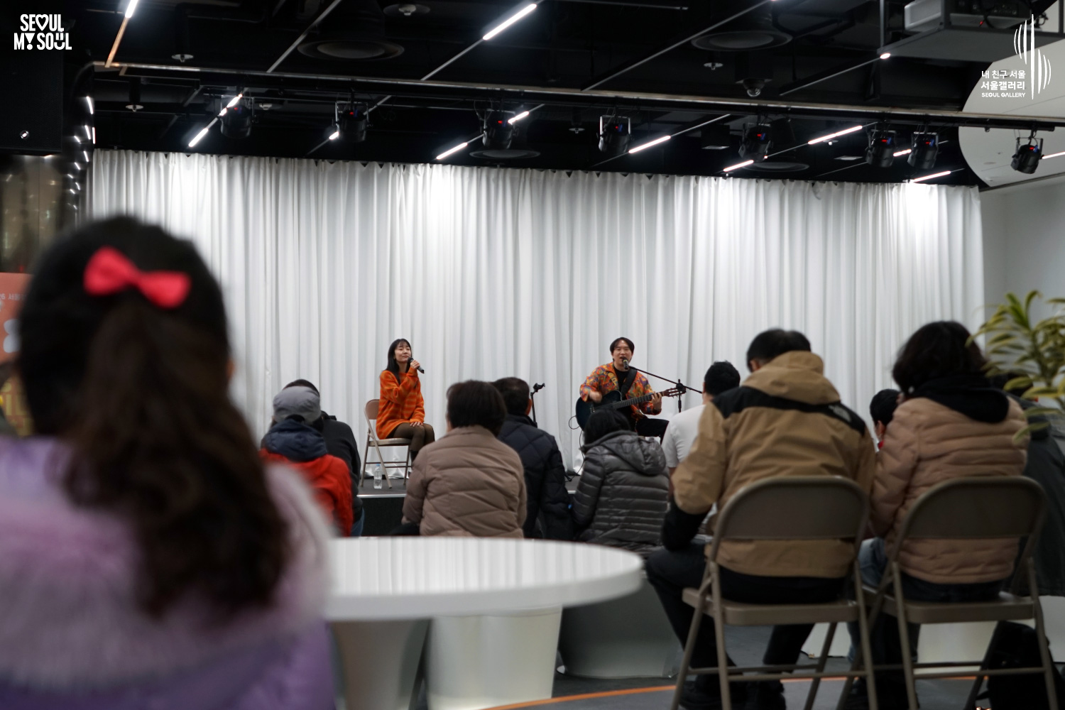 서울갤러리 무대에서 주황색 옷을 입은 여성 보컬이 마이크를 들고 노래하고, 옆에서 남성 연주자가 어쿠스틱 기타를 연주하는 어쿠스틱 듀오 공연을 객석의 관객들이 관람하고 있는 모습.