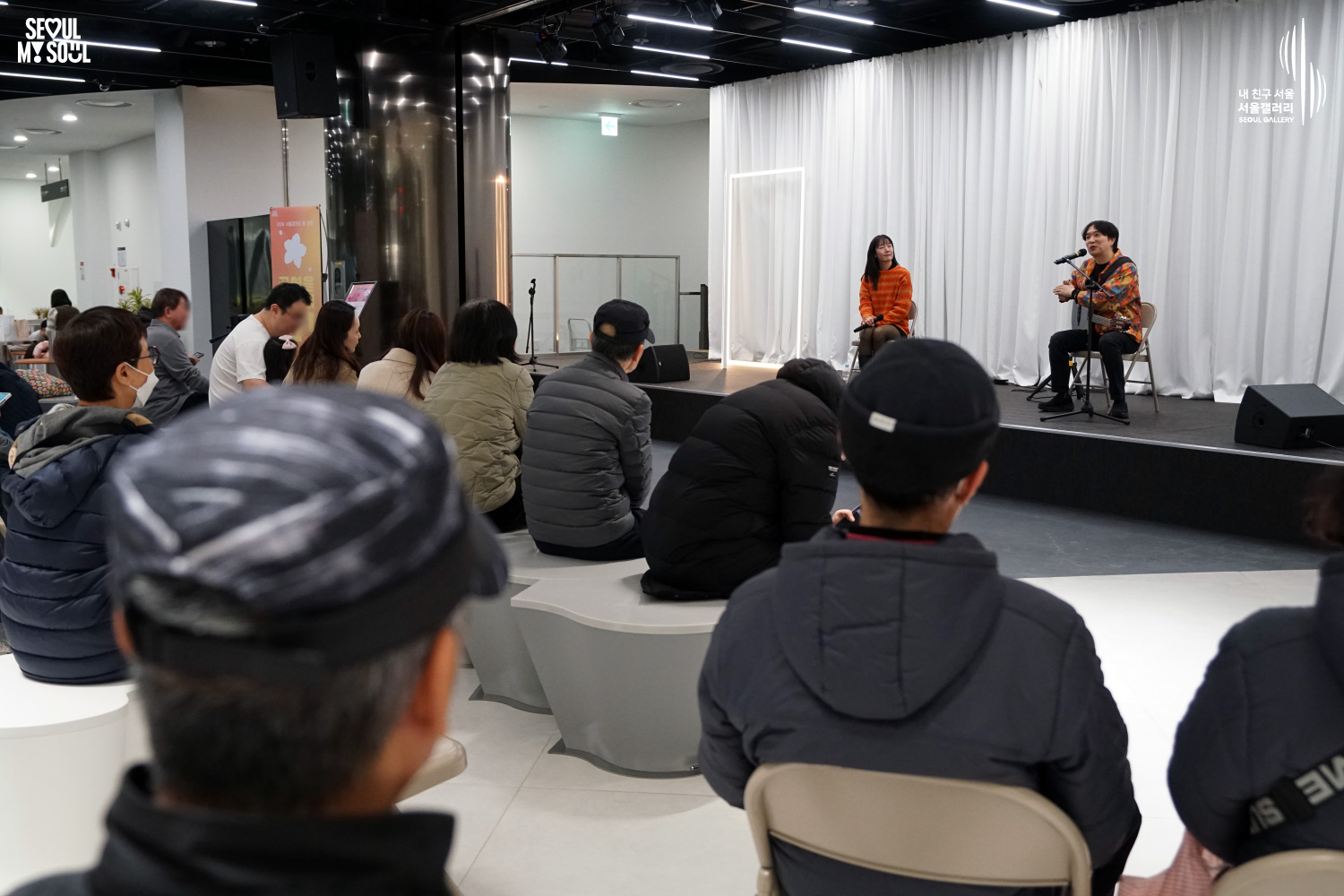 서울갤러리 무대에서 주황색 옷을 입은 여성 보컬이 마이크를 들고 노래하고, 옆에서 남성 연주자가 어쿠스틱 기타를 연주하는 어쿠스틱 듀오 공연을 객석의 관객들이 관람하고 있는 모습.