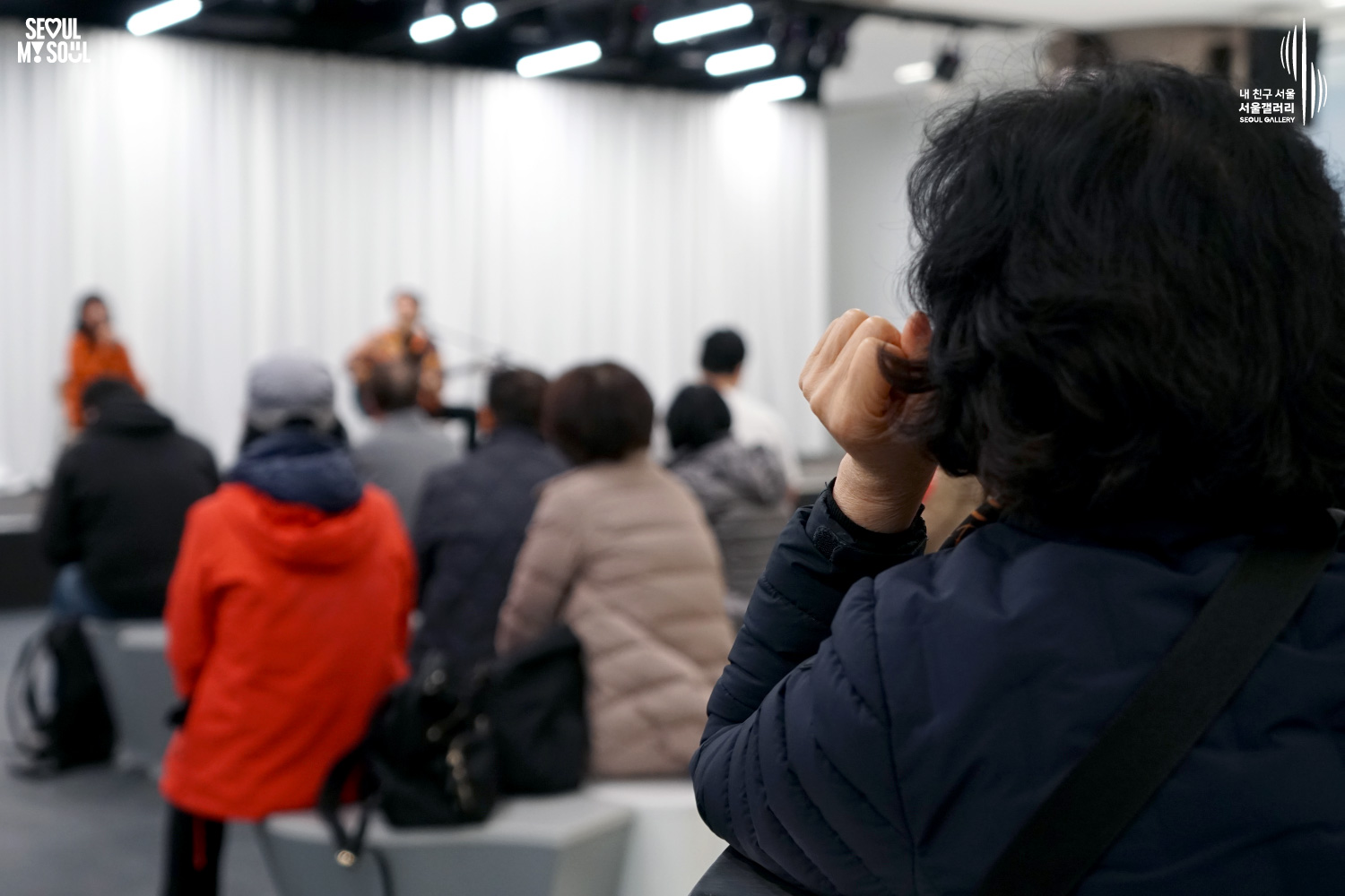 서울갤러리 무대에서 주황색 옷을 입은 여성 보컬이 마이크를 들고 노래하고, 옆에서 남성 연주자가 어쿠스틱 기타를 연주하는 어쿠스틱 듀오 공연을 객석의 관객들이 관람하고 있는 모습.