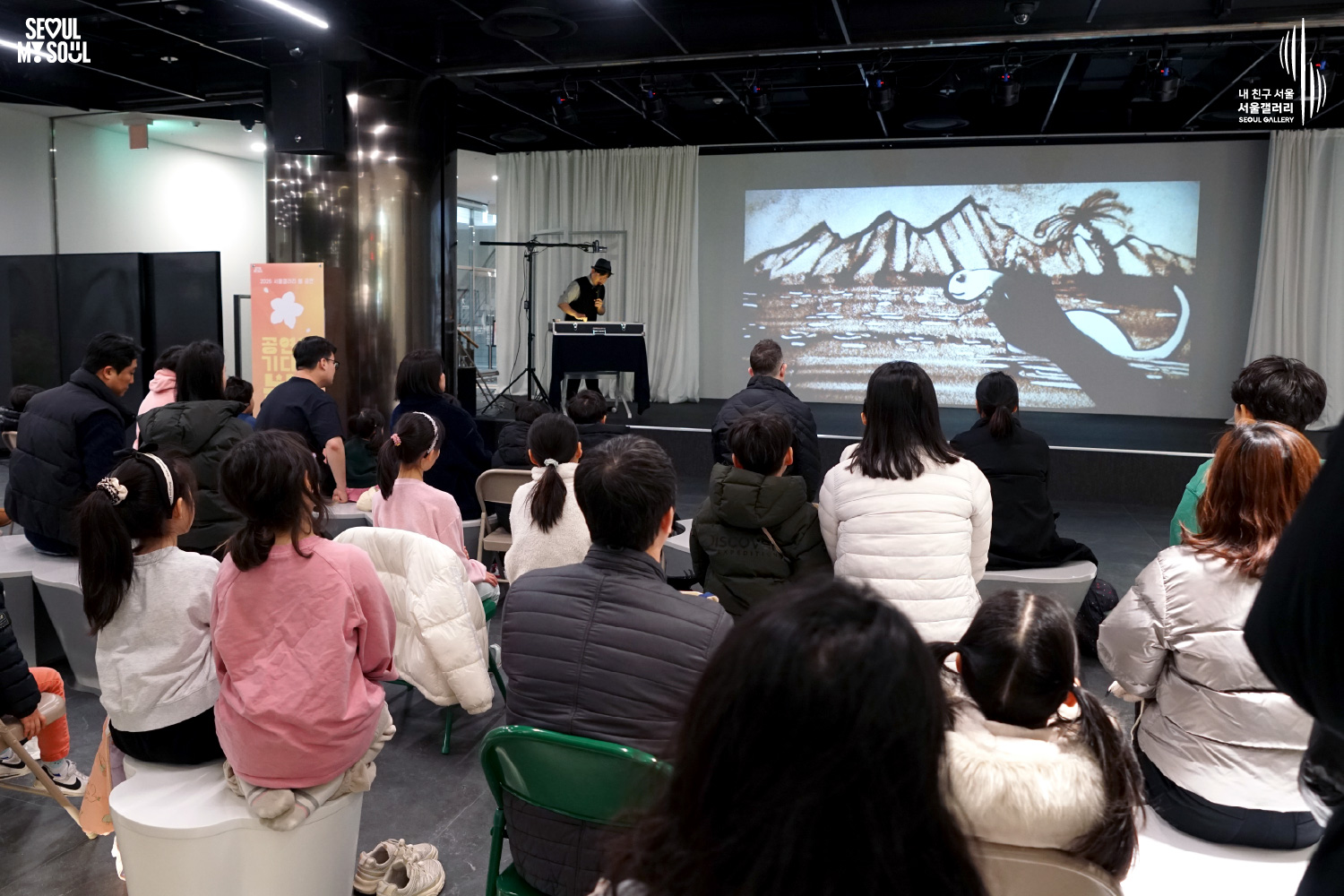 서울갤러리에서 진행된 샌드아트 공연 현장으로, 무대 위 작가가 모래로 그린 산과 물고기 모양 그림이 대형 스크린에 투사되고 있고 객석에는 어린이와 가족 관객들이 앉아 공연을 관람하고 있는 모습.