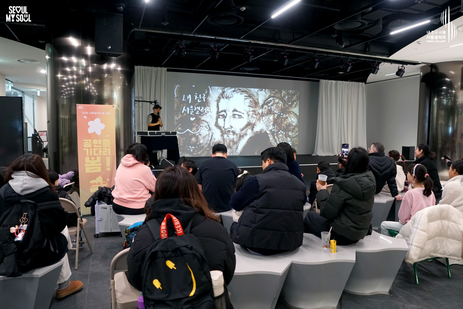 서울갤러리 '2026 서울갤러리 봄 공연 - 공연을 기다려 봄!' 행사에서 샌드아트 작가가 마이크를 들고 공연을 진행하고 있으며, 대형 스크린에는 '내 친구 서울갤러리' 글씨와 함께 인물 얼굴이 그려진 모래 그림이 투사되고 관객들이 관람하고 있는 모습.
