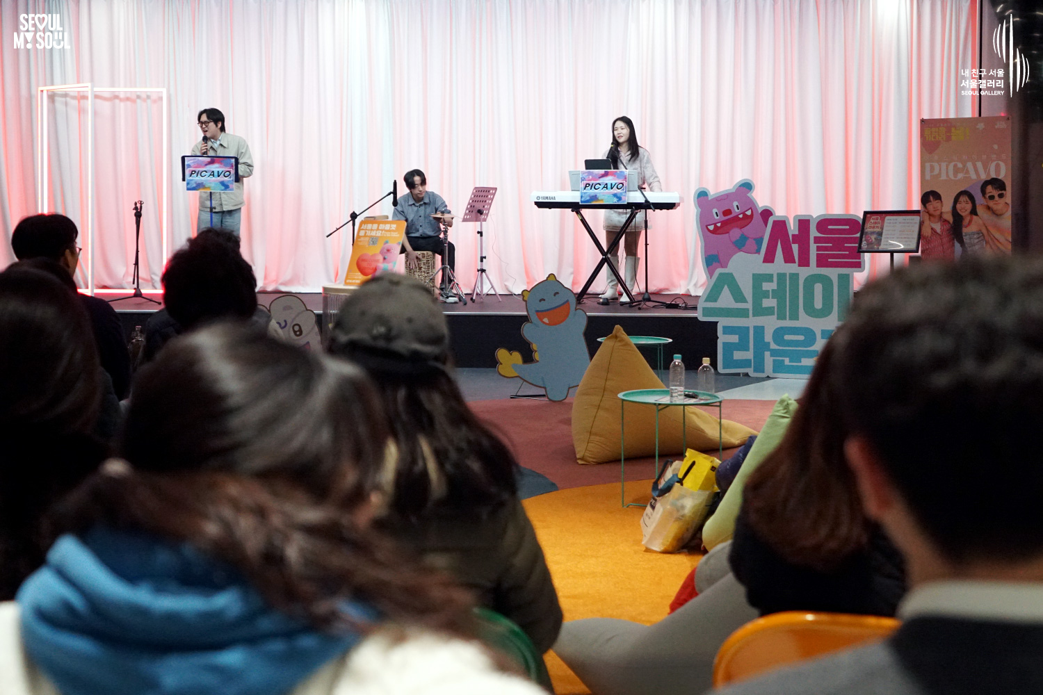 서울갤러리에서 열린 '서울 스테이 라운지' 공연 현장으로, 무대 위 가장 왼쪽 보컬이 마이크를 들고 노래하고 있으며 가운데에서 카혼 연주자가, 오른쪽에서 키보드 연주자가 공연을 펼치고 있고 객석에는 관객들이 앉아 관람하고 있는 모습.