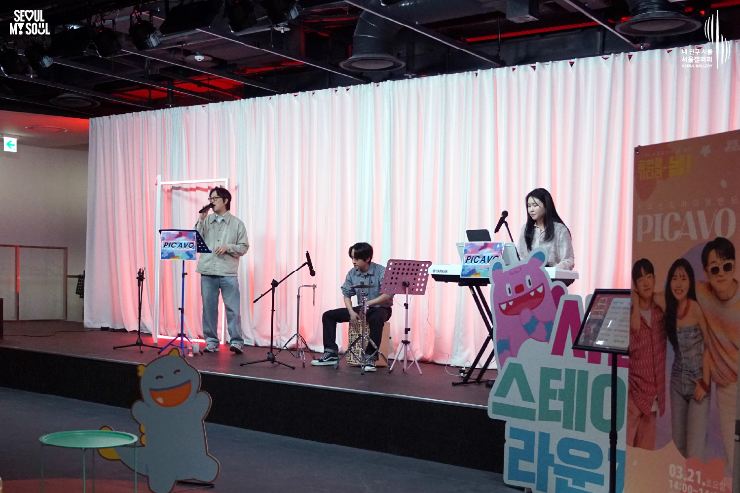 서울갤러리에서 열린 '서울 스테이 라운지' 공연 현장으로, 무대 위 가장 왼쪽 보컬이 마이크를 들고 노래하고 있으며 가운데에서 카혼 연주자가, 오른쪽에서 키보드 연주자가 공연을 펼치고 있는 모습