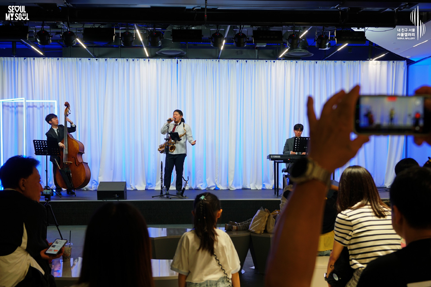 A jazz trio of double bass, saxophone, and Yamaha keyboard performs at Seoul Gallery before a seated audience, with "SEOUL MY SOUL" and "Seoul Gallery" logos visible.