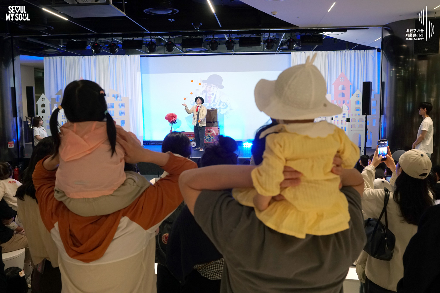 Children on their parents' shoulders watch a juggler perform on a stage with paper house decorations at Seoul Gallery, with "SEOUL MY SOUL" and "Seoul Gallery" logos visible.