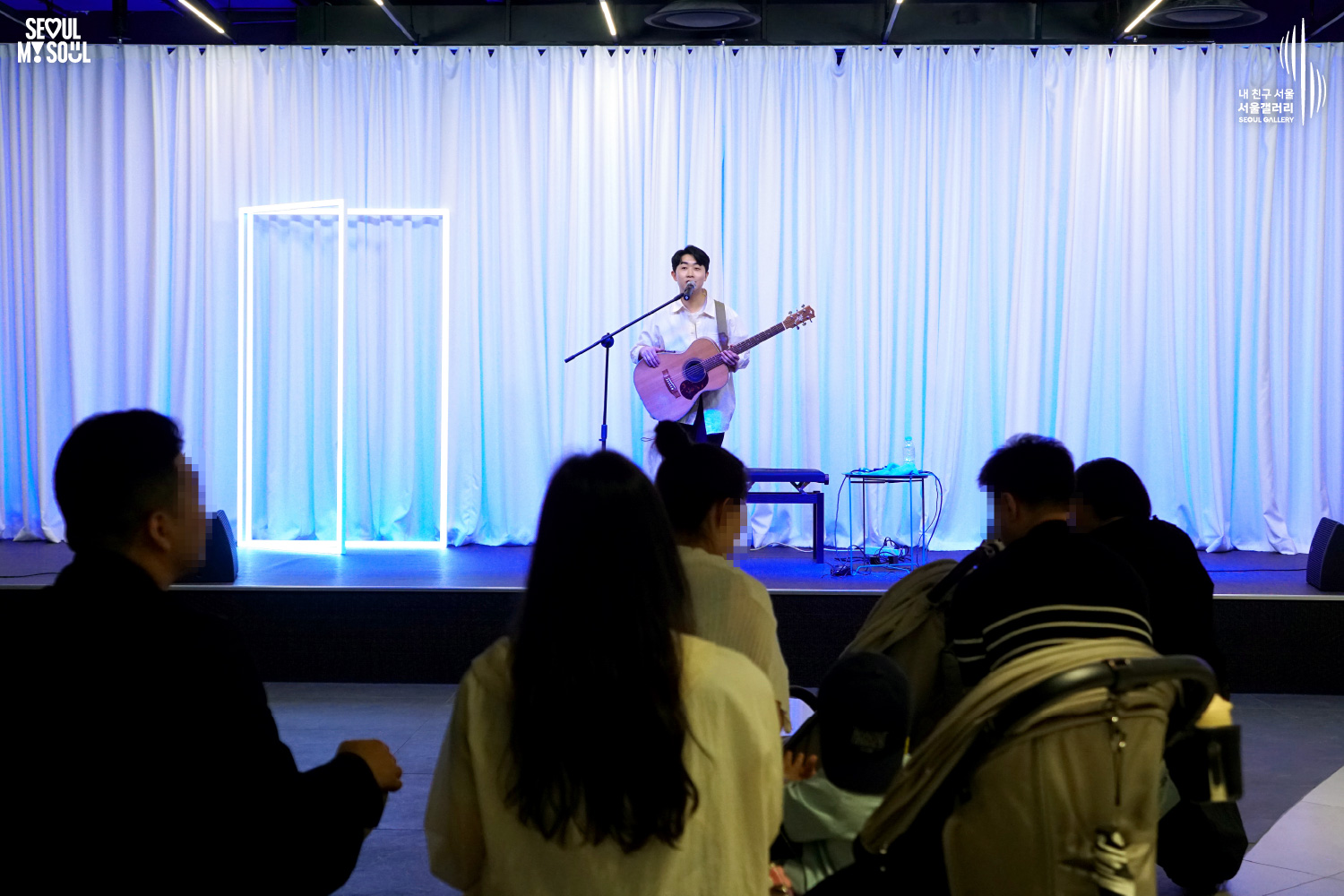 An image featuring the SEOUL MY SOUL logo and the My Friend Seoul - SEOUL GALLERY logo, capturing a live performance scene at the Seoul Gallery venue. On a stage set against a white curtain background with mixed blue and square neon lighting, a male singer-songwriter in a white shirt sings into a microphone with an acoustic guitar. A chair and pedal effectors are placed on the stage, and the shot captures the rear view of the audience looking toward the stage as they enjoy the performance.