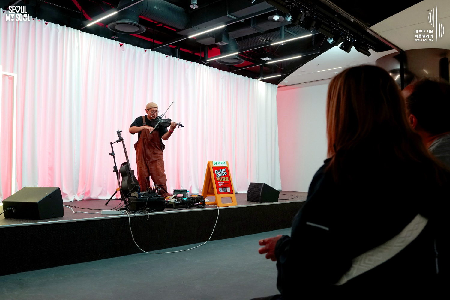 An image featuring the SEOUL MY SOUL logo and the My Friend Seoul - SEOUL GALLERY logo, capturing a live performance scene at the Seoul Gallery venue. On a stage against a white curtain background with pink lighting, the artist 'Guitar RoRo', wearing a beanie, glasses, a black T-shirt, and bright orange overalls, is immersed in playing an electric violin with the instrument on his shoulder and holding the bow. The stage includes an acoustic guitar on a stand, pedal effectors, speakers, a 'Guitar RoRo' sign, and an emergency exit sign, while the right foreground captures the rear view of an audience member looking toward the stage.