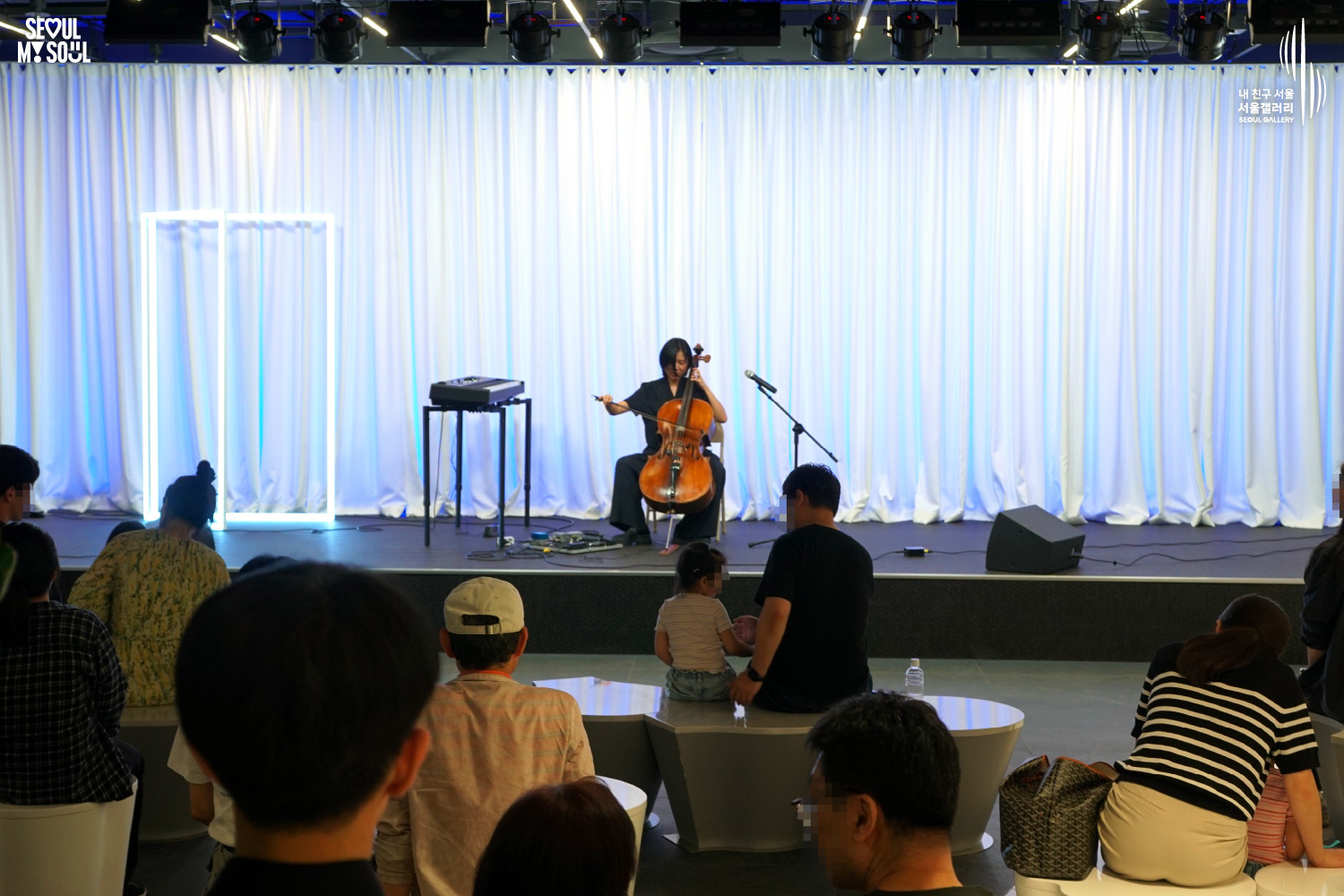 SEOUL MY SOUL 로고와 내 친구 서울 서울갤러리 SEOUL GALLERY 로고가 표기된 서울갤러리 공연장 사진으로, 푸른빛 조명이 비추는 흰색 커튼 배경의 무대 위에서 단발머리에 검은색 상의와 바지를 입은 'Big Violin Player 빅바이올린플레이어' 아티스트가 의자에 앉아 첼로를 연주하고 있으며, 무대 위에는 마이크 스탠드와 키보드, 스피커가 놓여 있고 객석에는 의자에 앉아 공연을 바라보고 있는 관람객들의 뒷모습이 함께 담긴 이미지입니다.