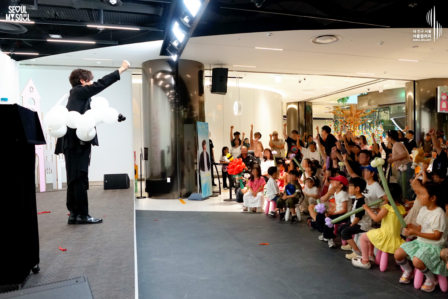 SEOUL MY SOUL 로고와 내 친구 서울 서울갤러리 SEOUL GALLERY 로고가 표기된 서울갤러리 지하 1층 공연장의 공연 현장 사진으로, 천장에 무대 조명이 설치된 실내 공간에서 중간 길이 머리의 재킷을 착용한 남성 아티스트가 무대 위에서 한 손을 높이 들어 공연을 진행하고 있으며, 객석의 어린이와 관람객들도 손을 들어 호응하고 있고 일부 어린이들은 풍선으로 만든 소품을 들고 있는 모습이 담긴 이미지입니다.