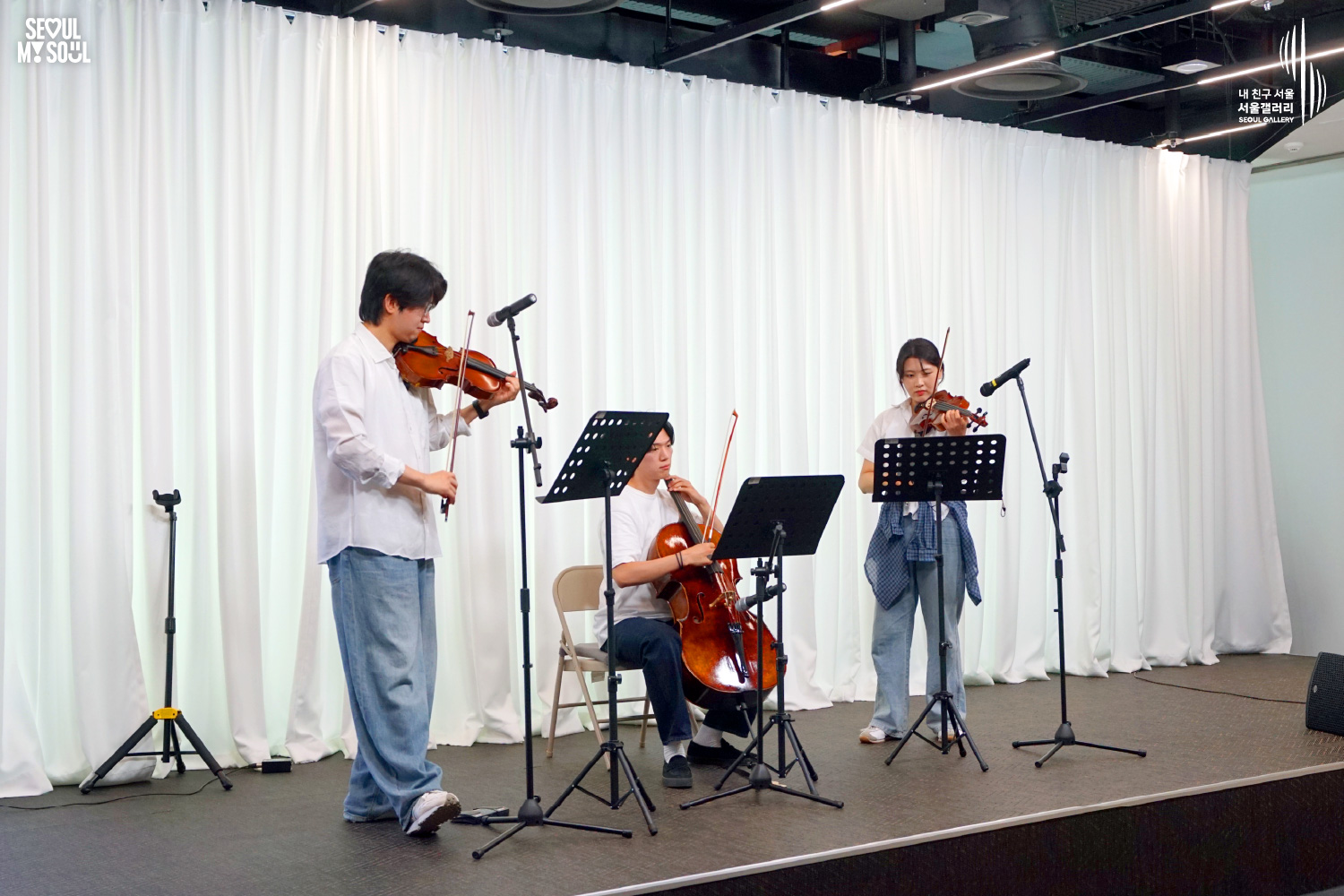 SEOUL MY SOUL 로고와 내 친구 서울 서울갤러리 SEOUL GALLERY 로고가 표기된 서울갤러리 공연장 무대 사진으로, 천장 조명이 비추는 흰색 커튼 배경의 무대 위에서 셔츠와 바지를 입은 남성 연주자 2명과 여성 연주자 1명이 바이올린과 첼로를 연주하며 집중하고 있으며, 무대 위에는 악보 거치대와 마이크 스탠드, 의자가 놓여 있는 모습이 담긴 이미지입니다.