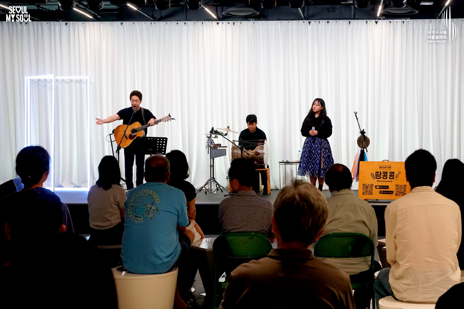 SEOUL MY SOUL 로고와 내 친구 서울 서울갤러리 SEOUL GALLERY 로고가 표기된 서울갤러리 공연장 사진으로, 천장 조명이 비추는 흰색 커튼 배경의 무대 위에서 기타를 멘 남성 싱어송라이터가 한쪽 팔을 뻗어 객석 방향을 가리키고 있으며, 타악기 앞에 앉은 남성 연주자와 마이크를 들고 서 있는 여성 싱어송라이터가 함께 무대에 서 있고, 객석에는 관람객들이 무대를 바라보고 있는 모습이 담긴 이미지입니다.
