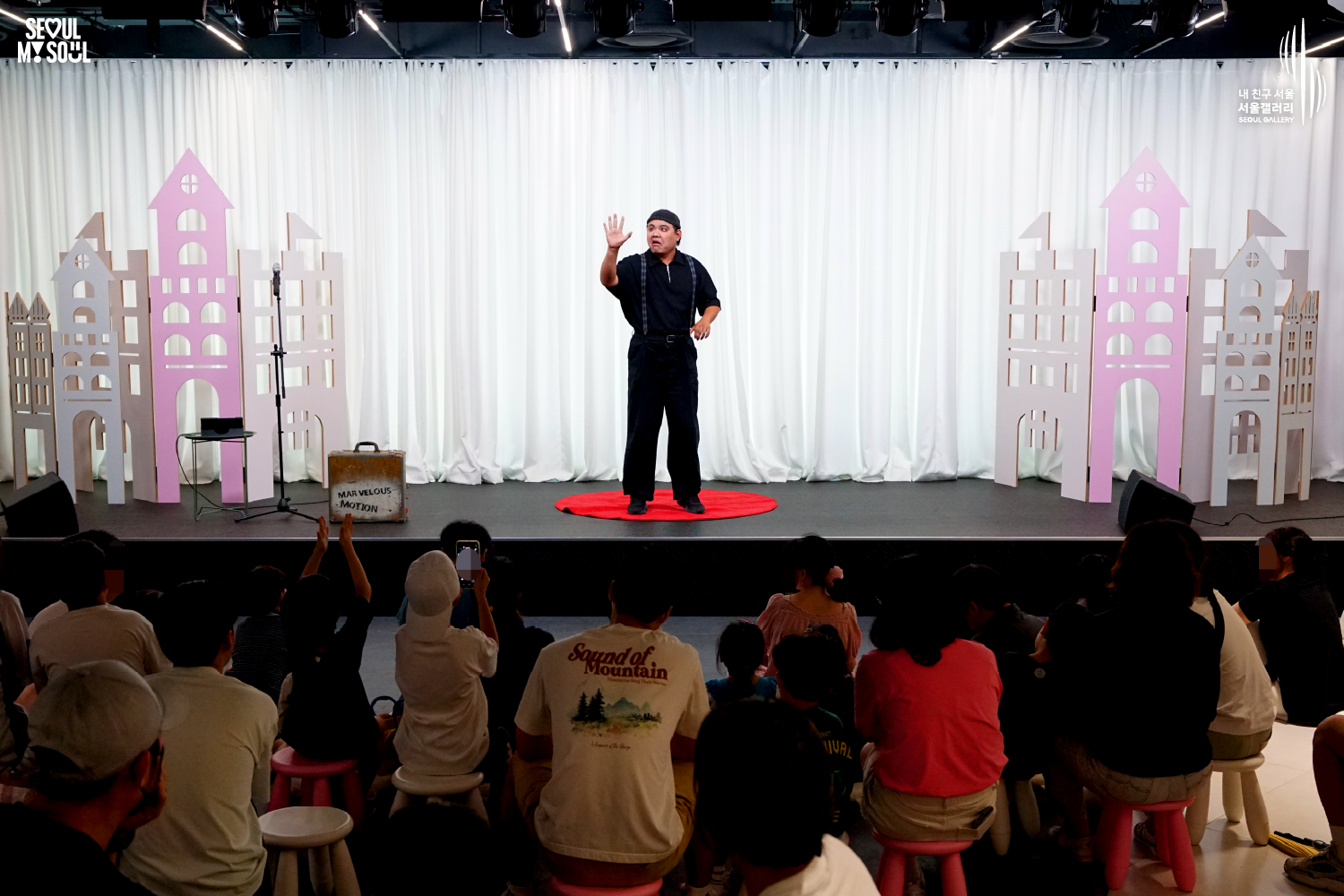 SEOUL MY SOUL 로고와 내 친구 서울 서울갤러리 SEOUL GALLERY 로고가 표기된 서울갤러리 공연장 사진으로, 천장에 무대 조명이 설치된 흰색 커튼 배경의 무대 위에서 모자를 착용한 남성 아티스트가 원형 무대 매트 위에 서서 한 손을 들어 공연을 진행하고 있으며, 무대 위에는 마이크 스탠드와 의자, 상자 형태의 공연 소품이 놓여 있고 객석에는 어린아이를 동반한 가족과 관람객들이 앉아 공연을 관람하고 있는 모습이 담긴 이미지입니다.