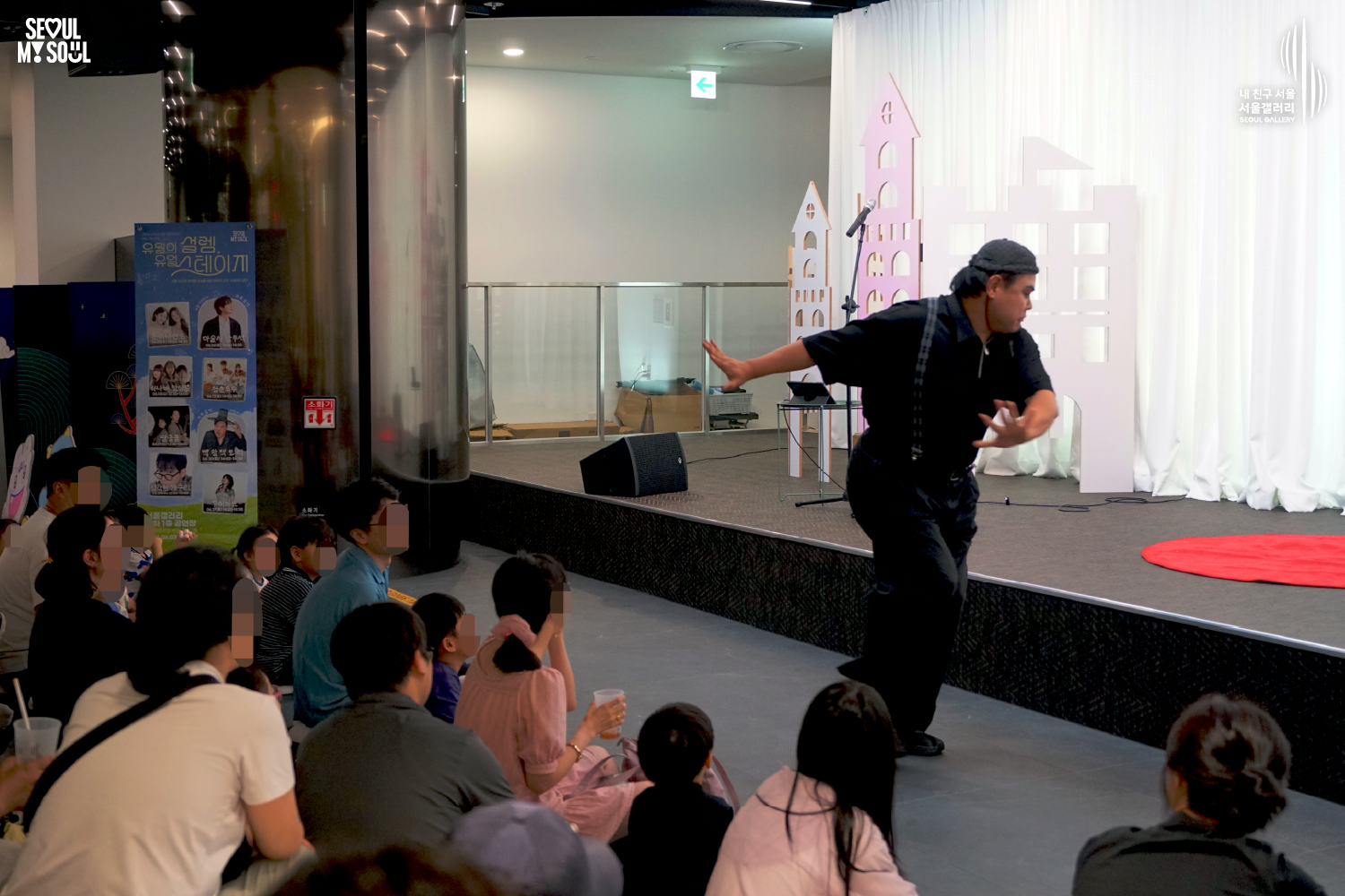 SEOUL MY SOUL 로고와 내 친구 서울 서울갤러리 SEOUL GALLERY 로고가 표기된 서울갤러리 공연장 사진으로, 천장에 무대 조명이 설치된 실내 공간에서 모자를 착용한 남성 아티스트가 무대 앞 객석 가까이에서 양팔을 펼친 채 공연을 진행하고 있으며, 무대 옆에는 공연 안내 배너와 마이크 스탠드, 스피커가 놓여 있고 객석 앞줄의 어린이 관람객들이 무대를 바라보고 있는 모습이 담긴 이미지입니다.