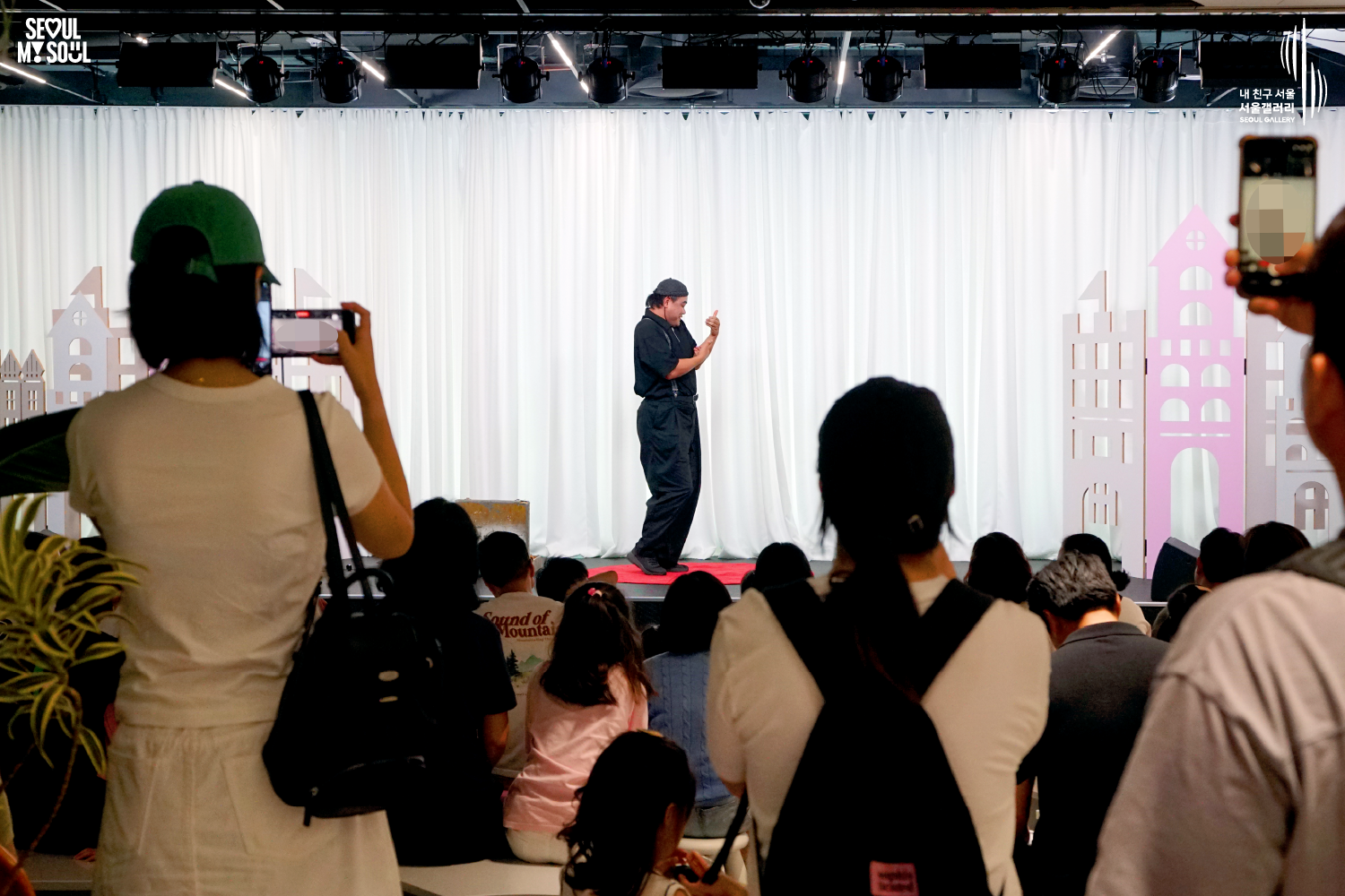 SEOUL MY SOUL 로고와 내 친구 서울 서울갤러리 SEOUL GALLERY 로고가 표기된 서울갤러리 공연장 사진으로, 천장에 무대 조명이 설치된 흰색 커튼 배경의 무대 위에서 모자를 착용한 남성 아티스트가 원형 무대 매트 위에 서서 양손을 모아 공연을 진행하고 있으며, 객석에는 관람객들이 앉아 공연을 관람하고 있고 일부 관람객은 휴대전화로 무대를 촬영하고 있는 모습이 담긴 이미지입니다.