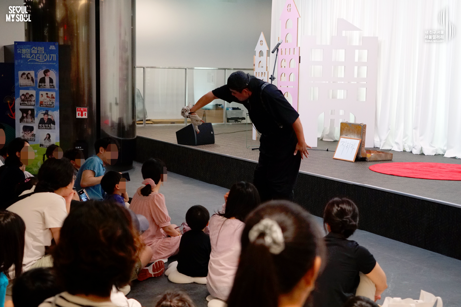 SEOUL MY SOUL 로고와 내 친구 서울 서울갤러리 SEOUL GALLERY 로고가 표기된 서울갤러리 공연장 사진으로, 천장에 무대 조명이 설치된 실내 공간에서 모자를 착용한 남성 아티스트가 손에 든 인형 형태의 공연 소품을 관객들에게 보여주며 공연을 진행하고 있으며, 무대 위에는 마이크 스탠드와 상자, 안내판이 놓여 있고 객석의 어린이와 관람객들이 무대를 바라보고 있는 모습이 담긴 이미지입니다.