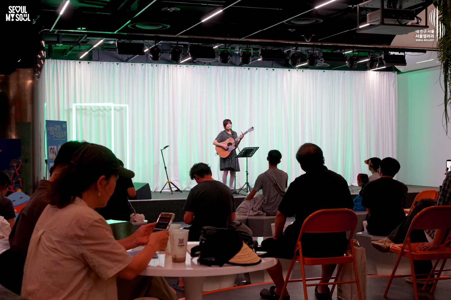 SEOUL MY SOUL 로고와 내 친구 서울 서울갤러리 SEOUL GALLERY 로고가 표기된 서울갤러리 공연장 사진으로, 연둣빛 조명과 사각 네온 조명이 어우러진 흰색 커튼 배경의 무대 위에서 단발머리에 원피스를 입은 여성 싱어송라이터가 어쿠스틱 기타를 메고 마이크 앞에서 노래를 부르고 있으며, 무대 위에는 마이크 스탠드와 악보 거치대, 모니터 스피커, 공연 안내판이 놓여 있고 객석에는 다양한 연령대의 관람객들이 공연을 관람하고 있는 모습이 담긴 이미지입니다.