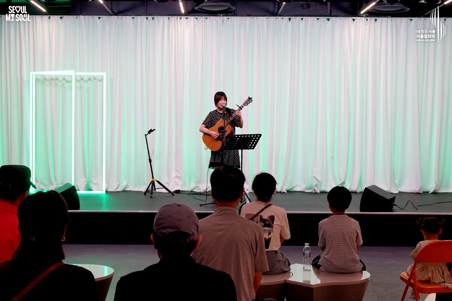 SEOUL MY SOUL 로고와 내 친구 서울 서울갤러리 SEOUL GALLERY 로고가 표기된 서울갤러리 공연장 사진으로, 연둣빛 조명과 사각 네온 조명이 어우러진 흰색 커튼 배경의 무대 위에서 단발머리에 원피스를 입은 여성 싱어송라이터가 어쿠스틱 기타를 메고 마이크 앞에서 노래를 부르고 있으며, 무대 위에는 마이크 스탠드와 악보 거치대, 모니터 스피커가 놓여 있고 객석에는 어린이를 포함한 다양한 연령대의 관람객들이 공연을 관람하고 있는 모습이 담긴 이미지입니다.