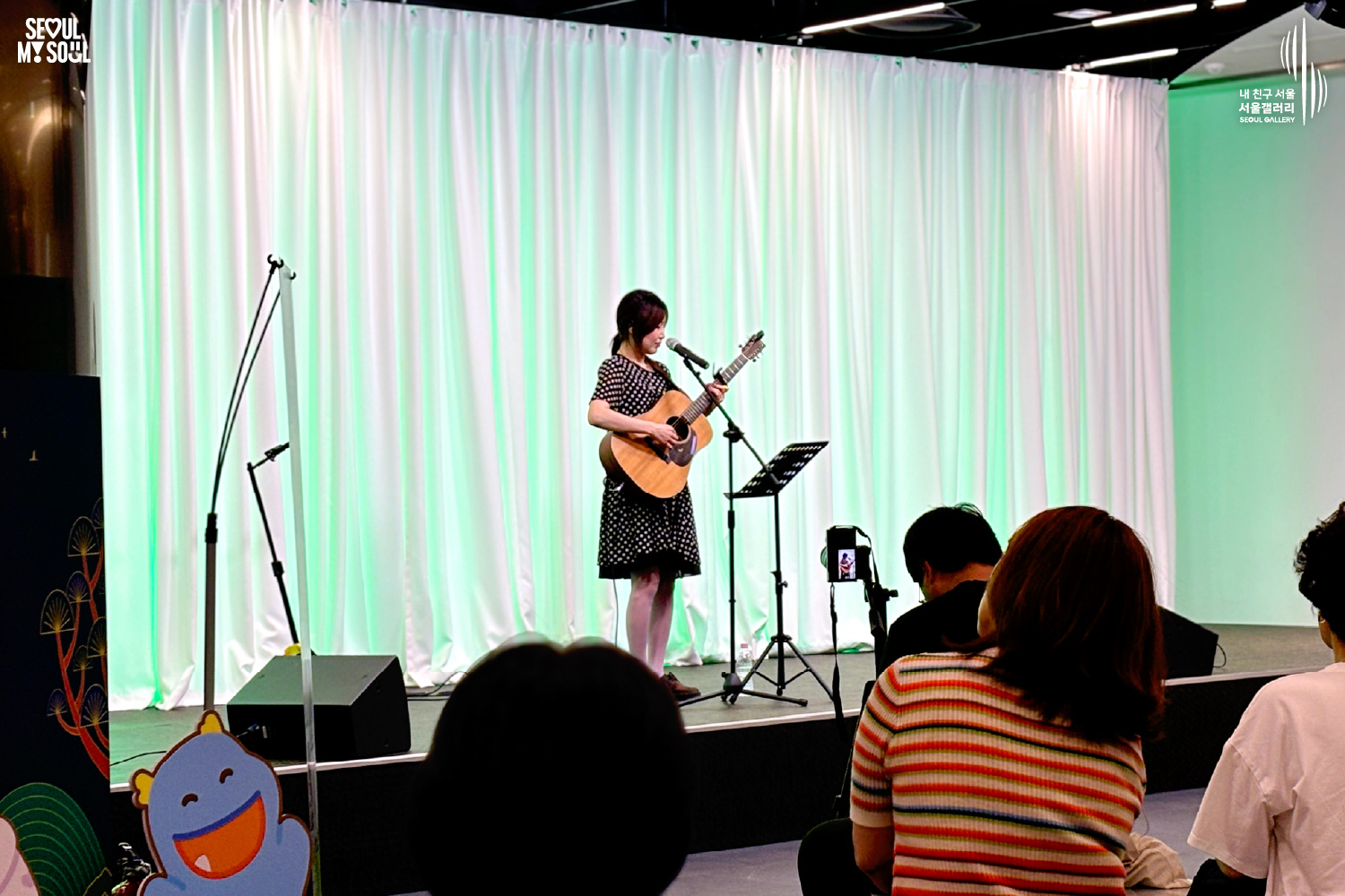 SEOUL MY SOUL 로고와 내 친구 서울 서울갤러리 SEOUL GALLERY 로고가 표기된 서울갤러리 공연장 사진으로, 연둣빛 조명과 사각 네온 조명이 어우러진 흰색 커튼 배경의 무대 위에서 단발머리에 원피스를 입은 여성 싱어송라이터가 어쿠스틱 기타를 메고 마이크 앞에서 노래를 부르고 있으며, 무대 위에는 마이크 스탠드와 악보 거치대, 모니터 스피커가 놓여 있고 객석에는 다양한 연령대의 관람객들이 공연을 관람하며 일부는 휴대전화로 무대를 촬영하고 있는 모습이 담긴 이미지입니다.