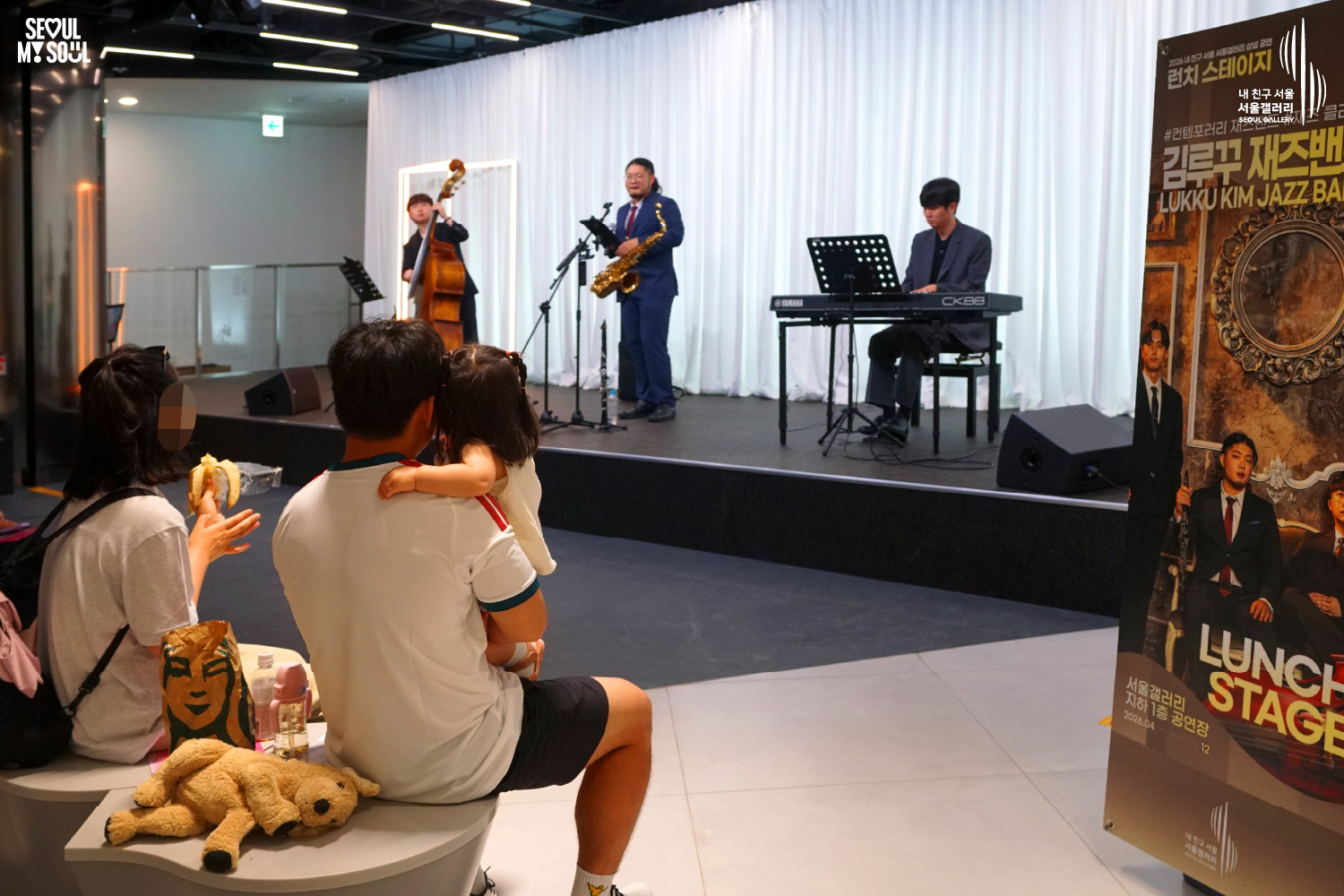 SEOUL MY SOUL 로고와 내 친구 서울 서울갤러리 SEOUL GALLERY 로고가 표기된 서울갤러리 공연장 사진으로, 흰색 커튼 배경의 무대 위에서 묶은 머리에 안경을 착용한 남성 연주자가 마이크 앞에서 노래를 부르고 있으며, 다른 남성 연주자는 콘트라베이스를 연주하고 재킷을 착용한 남성 연주자는 키보드를 연주하고 있다. 무대 위에는 색소폰과 마이크 스탠드, 키보드, 공연 안내판이 놓여 있고 객석에는 어린아이를 안은 가족을 포함한 관람객들이 공연을 관람하고 있는 모습이 담긴 이미지입니다.