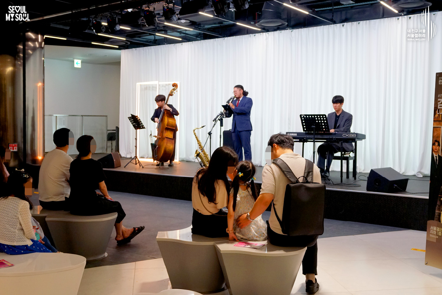 SEOUL MY SOUL 로고와 내 친구 서울 서울갤러리 SEOUL GALLERY 로고가 표기된 서울갤러리 공연장 사진으로, 흰색 커튼 배경의 무대 위에서 재킷을 착용한 남성 연주자가 키보드를 연주하고, 묶은 머리에 안경을 착용한 남성 연주자가 마이크 앞에서 노래를 부르고 있으며, 다른 남성 연주자가 콘트라베이스를 연주하고 있다. 무대 위에는 키보드와 콘트라베이스, 색소폰, 마이크 스탠드, 악보 거치대, 모니터 스피커가 놓여 있고 객석에는 어린이를 포함한 다양한 연령대의 관람객들이 공연을 관람하고 있는 모습이 담긴 이미지입니다.