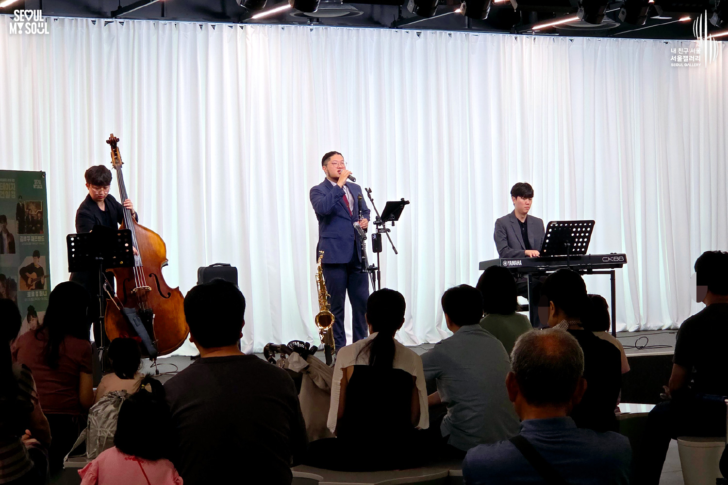 SEOUL MY SOUL 로고와 내 친구 서울 서울갤러리 SEOUL GALLERY 로고가 표기된 서울갤러리 공연장 사진으로, 흰색 커튼 배경의 무대 위에서 묶은 머리에 안경을 착용한 남성 연주자가 마이크를 들고 있으며, 다른 남성 연주자는 콘트라베이스를 연주하고 재킷을 착용한 남성 연주자는 키보드를 연주하고 있다. 무대 위에는 색소폰과 마이크 스탠드, 악보 거치대, 키보드가 놓여 있고 객석에는 다양한 연령대의 관람객들이 공연을 관람하고 있는 모습이 담긴 이미지입니다.