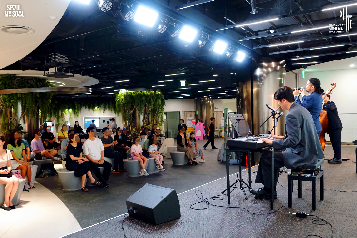 SEOUL MY SOUL 로고와 내 친구 서울 서울갤러리 SEOUL GALLERY 로고가 표기된 서울갤러리 지하 1층 공연장의 공연 현장 사진으로, 천장에 무대 조명이 설치된 실내 공간에서 재킷을 착용한 남성 연주자가 키보드를 연주하고, 묶은 머리에 안경을 착용한 남성 연주자가 마이크 앞에서 노래를 부르고 있으며, 다른 남성 연주자가 콘트라베이스를 연주하고 있다. 무대 위에는 키보드와 마이크 스탠드, 콘트라베이스, 색소폰, 모니터 스피커가 놓여 있고 다양한 연령대의 관람객들이 공연을 관람하고 있는 모습이 담긴 이미지입니다.