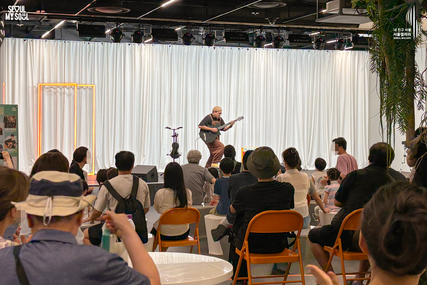 SEOUL MY SOUL 로고와 내 친구 서울 서울갤러리 SEOUL GALLERY 로고가 표기된 서울갤러리 공연장 사진으로, 흰색 커튼 배경의 무대 위에서 비니 모자와 안경을 착용한 멜빵 의상의 남성 연주자가 기타를 연주하며 집중하고 있으며, 무대 위에는 마이크 스탠드와 기타 거치대, 공연 안내판이 놓여 있고 객석에는 다양한 연령대의 관람객들이 공연을 관람하고 있는 모습이 담긴 이미지입니다.