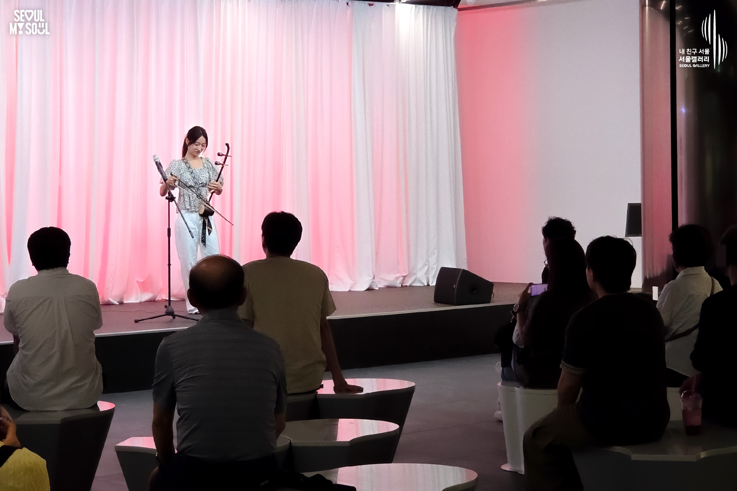 SEOUL MY SOUL 로고와 내 친구 서울 서울갤러리 SEOUL GALLERY 로고가 표기된 서울갤러리 공연장 사진으로, 분홍빛 조명이 비추는 흰색 커튼 배경의 무대 위에서 긴 머리를 묶은 여성 연주자 우하린이 해금을 연주하기 위해 악기를 조정하고 있으며, 무대 위에는 마이크 스탠드와 모니터 스피커가 놓여 있고 객석에는 다양한 연령대의 관람객들이 공연을 관람하고 있는 모습이 담긴 이미지입니다.