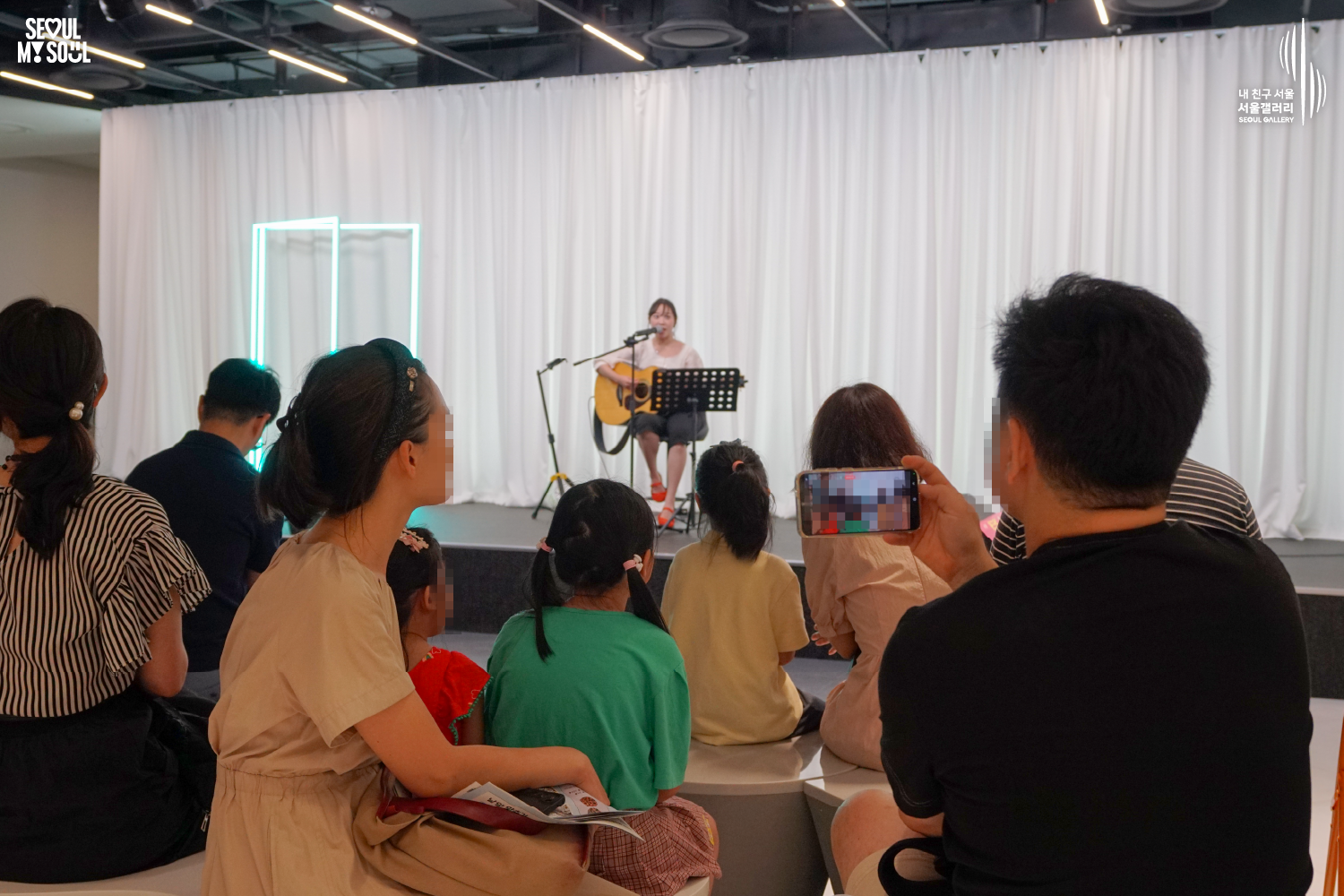 SEOUL MY SOUL 로고와 내 친구 서울 서울갤러리 SEOUL GALLERY 로고가 표기된 서울갤러리 공연장 사진으로, 흰색 커튼과 청록빛 사각 네온 조명이 보이는 무대 위에서 긴 머리를 묶은 여성 싱어송라이터가 의자에 앉아 어쿠스틱 기타를 연주하며 마이크 앞에서 노래를 부르고 있으며, 객석에는 어린이를 포함한 다양한 연령대의 관람객들이 공연을 관람하고 있고 우측 전경의 관람객은 휴대전화로 무대를 촬영하고 있는 모습이 담긴 이미지입니다.