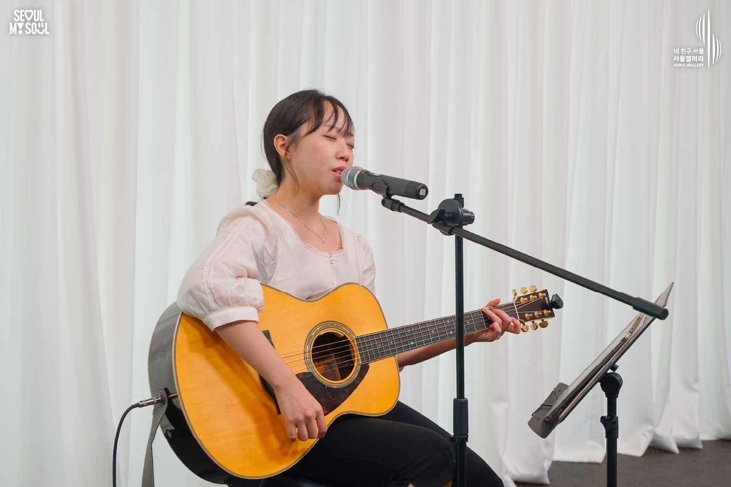SEOUL MY SOUL 로고와 내 친구 서울 서울갤러리 SEOUL GALLERY 로고가 표기된 서울갤러리 공연장 무대 사진으로, 은은한 조명이 비추는 흰색 커튼 배경의 무대 위에서 긴 머리를 묶은 여성 싱어송라이터가 의자에 앉아 어쿠스틱 기타를 연주하며 눈을 감고 마이크 앞에서 노래를 부르고 있으며, 앞쪽에는 마이크 스탠드와 보면대가 놓여 있는 모습이 담긴 이미지입니다.