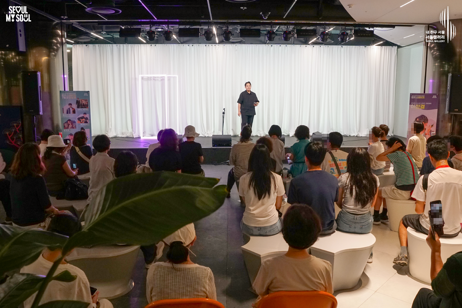 SEOUL MY SOUL 로고와 내 친구 서울 서울갤러리 SEOUL GALLERY 로고가 표기된 서울갤러리 공연장 사진으로, 천장에 무대 조명이 설치되고 흰색 커튼과 보랏빛 사각 네온 조명이 보이는 무대 위에서 짧은 머리의 남성 연주자가 관객을 바라보며 서 있고, 무대 위에는 마이크 스탠드와 모니터 스피커, 공연 안내판이 놓여 있으며 객석에는 다양한 연령대의 관람객들이 앉아 무대를 바라보고 있는 모습이 담긴 이미지입니다.