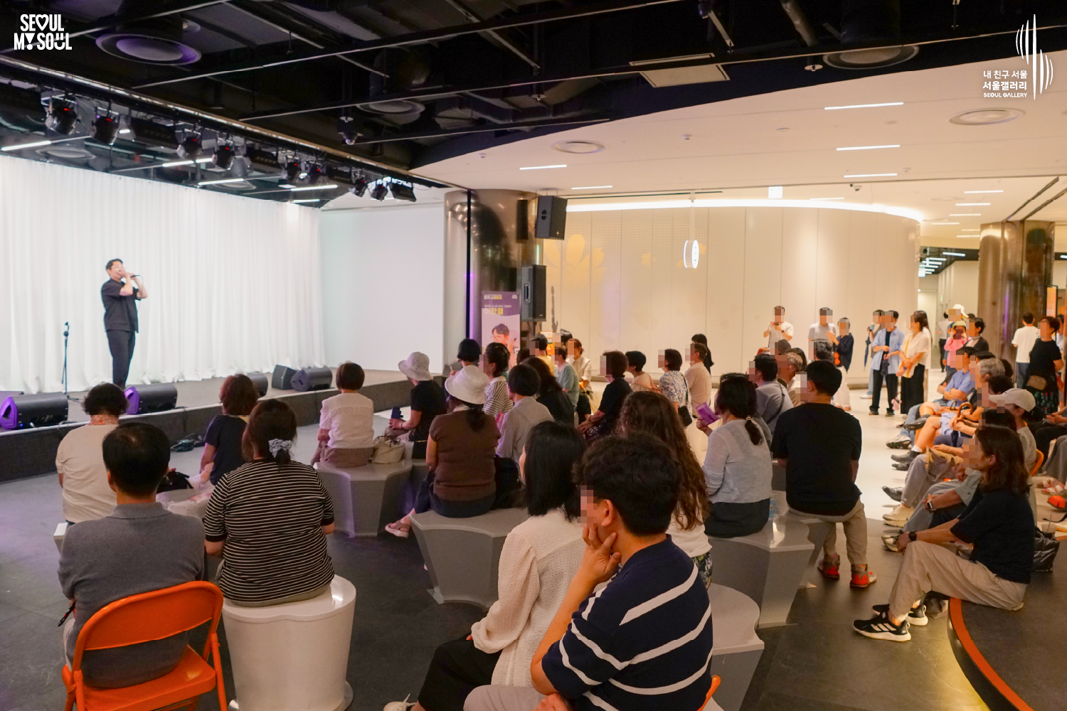 SEOUL MY SOUL 로고와 내 친구 서울 서울갤러리 SEOUL GALLERY 로고가 표기된 서울갤러리 지하 1층 공연장의 공연 현장 사진으로, 천장에 무대 조명이 설치되고 흰색 커튼을 배경으로 한 무대 위에서 짧은 머리의 남성 연주자가 하모니카를 두 손으로 들고 연주하고 있으며, 무대 앞에는 모니터 스피커가 놓여 있고 객석과 통로에는 다양한 연령대의 관람객들이 앉거나 서서 공연을 관람하고 있는 모습이 담긴 이미지입니다.