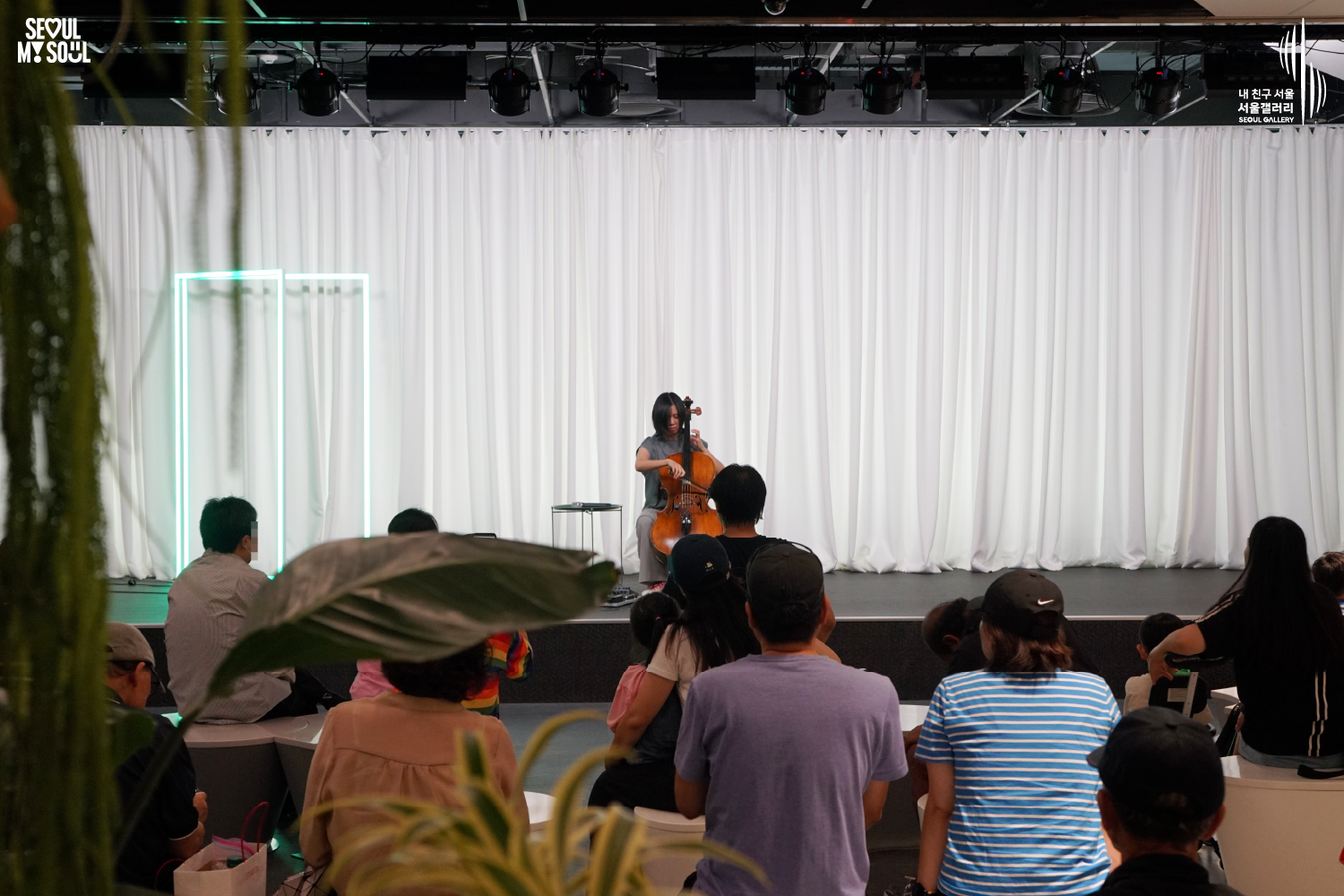SEOUL MY SOUL 로고와 내 친구 서울 서울갤러리 SEOUL GALLERY 로고가 표기된 서울갤러리 공연장 사진으로, 흰색 커튼과 청록빛 사각 네온 조명이 보이는 무대 위에서 단발머리의 여성 연주자가 의자에 앉아 첼로를 활로 연주하고 있으며, 무대 위에는 작은 테이블과 페달 이펙터가 놓여 있고 객석에는 다양한 연령대의 관람객들이 앉아 공연을 관람하고 있는 모습이 담긴 이미지입니다.