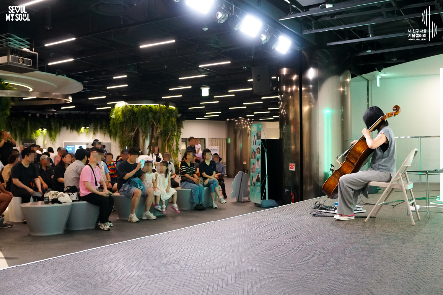 SEOUL MY SOUL 로고와 내 친구 서울 서울갤러리 SEOUL GALLERY 로고가 표기된 서울갤러리 지하 1층 공연장의 공연 현장 사진으로, 천장에 무대 조명이 설치된 실내 공간에서 단발머리의 여성 연주자가 의자에 앉아 첼로를 활로 연주하고 있으며, 무대 위에는 페달 이펙터와 의자가 놓여 있고 객석에는 어린이를 포함한 다양한 연령대의 관람객들이 앉아 공연을 관람하고 있는 모습이 담긴 이미지입니다.