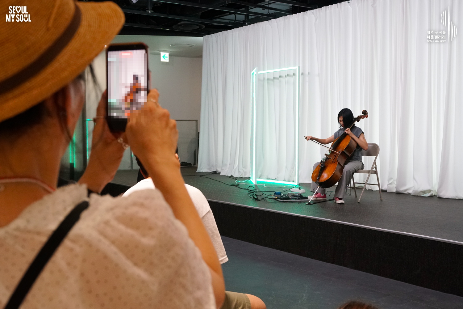 SEOUL MY SOUL 로고와 내 친구 서울 서울갤러리 SEOUL GALLERY 로고가 표기된 서울갤러리 공연장 사진으로, 은은한 조명이 비추는 흰색 커튼과 청록빛 사각 네온 조명을 배경으로 단발머리의 여성 연주자가 의자에 앉아 첼로를 활로 연주하고 있으며, 무대 위에는 페달 이펙터와 케이블이 놓여 있고 좌측 전경의 관람객이 휴대전화로 연주 모습을 촬영하고 있는 모습이 담긴 이미지입니다.