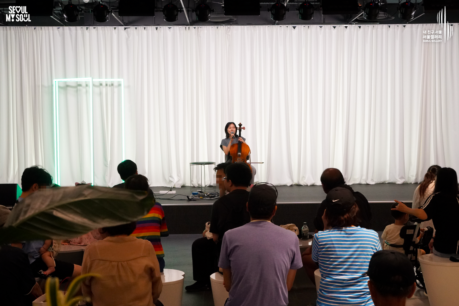 SEOUL MY SOUL 로고와 내 친구 서울 서울갤러리 SEOUL GALLERY 로고가 표기된 서울갤러리 공연장 사진으로, 흰색 커튼과 청록빛 사각 네온 조명이 보이는 무대 위에서 단발머리의 여성 연주자가 의자에 앉아 첼로를 세운 채 마이크를 들고 관객을 바라보며 이야기하고 있으며, 무대 위에는 작은 테이블과 페달 이펙터가 놓여 있고 객석에는 다양한 연령대의 관람객들이 공연을 관람하고 있는 모습이 담긴 이미지입니다.