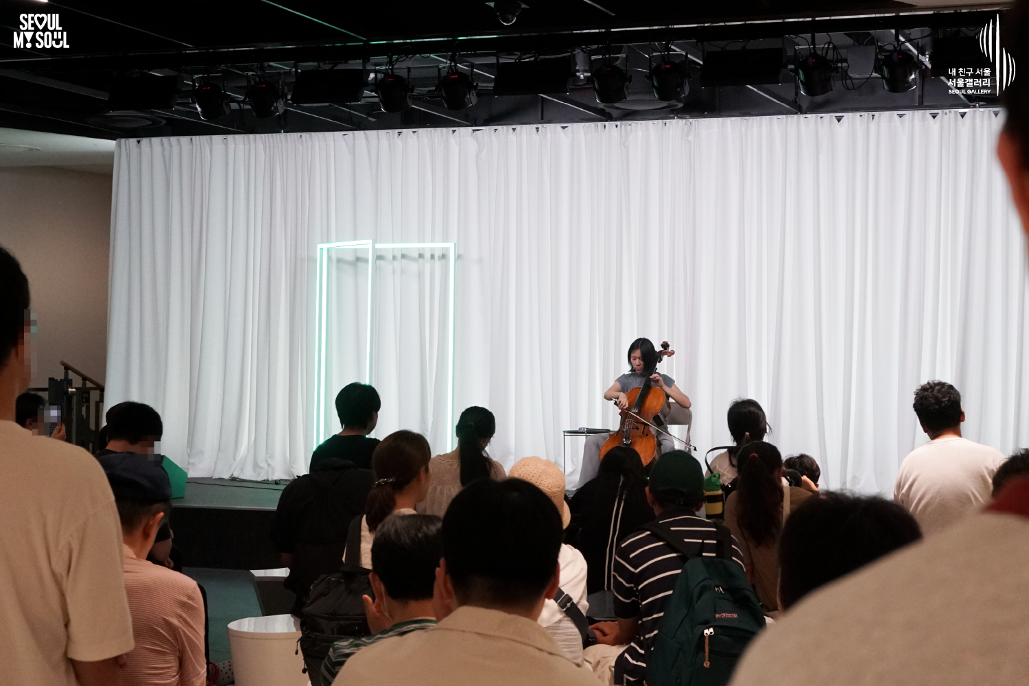 SEOUL MY SOUL 로고와 내 친구 서울 서울갤러리 SEOUL GALLERY 로고가 표기된 서울갤러리 공연장 사진으로, 천장에 무대 조명이 설치되고 흰색 커튼과 청록빛 사각 네온 조명이 보이는 무대 위에서 단발머리의 여성 연주자가 의자에 앉아 첼로를 활로 연주하고 있으며, 전경에는 다양한 연령대의 관람객들이 앉거나 서서 무대를 바라보며 공연을 관람하고 있는 모습이 담긴 이미지입니다.