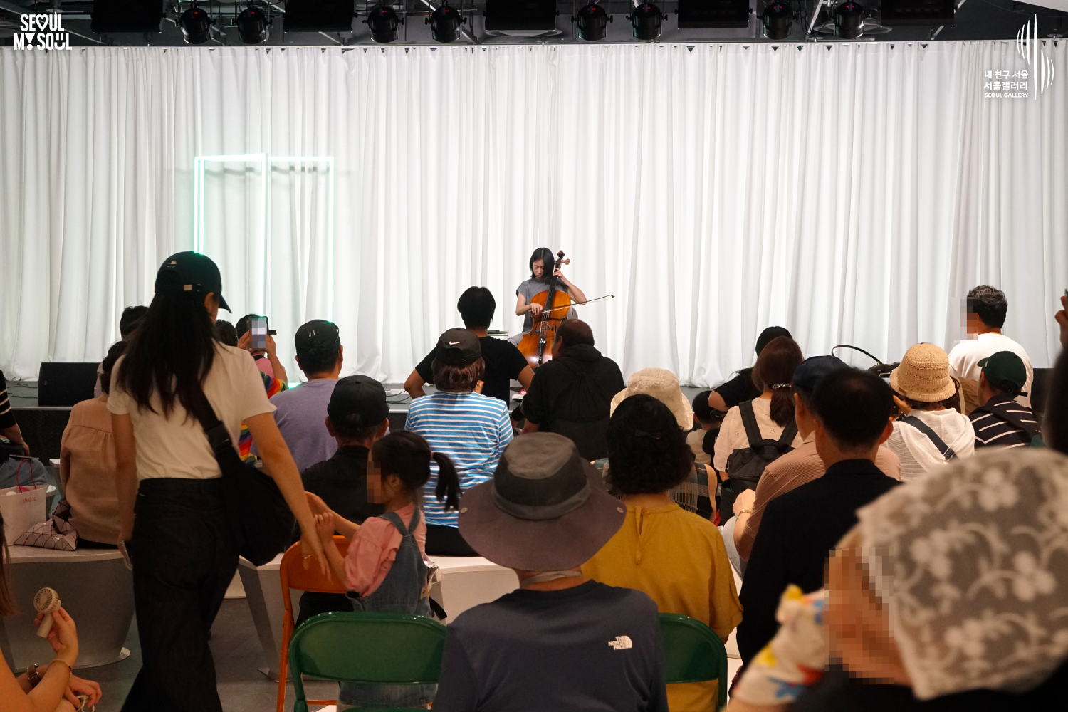 SEOUL MY SOUL 로고와 내 친구 서울 서울갤러리 SEOUL GALLERY 로고가 표기된 서울갤러리 공연장 사진으로, 천장에 무대 조명이 설치되고 흰색 커튼과 청록빛 사각 네온 조명이 보이는 무대 위에서 단발머리의 여성 연주자가 의자에 앉아 첼로를 활로 연주하고 있으며, 객석에는 어린이를 포함한 다양한 연령대의 관람객들이 앉아 공연을 관람하고 일부 관람객은 휴대전화로 무대를 촬영하고 있는 모습이 담긴 이미지입니다.