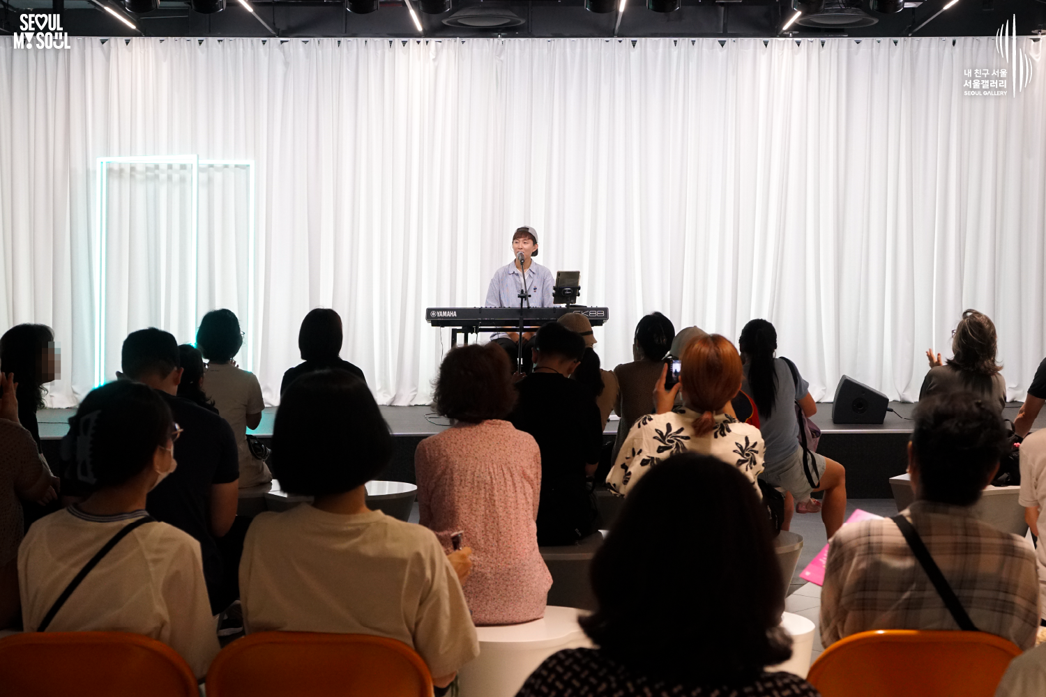 SEOUL MY SOUL 로고와 내 친구 서울 서울갤러리 SEOUL GALLERY 로고가 표기된 서울갤러리 공연장 사진으로, 흰색 커튼과 청록빛 사각 네온 조명이 보이는 무대 위에서 모자를 뒤로 쓴 짧은 머리의 남성 연주자가 의자에 앉아 키보드를 연주하며 마이크 앞에서 노래를 부르고 있으며, 객석에는 다양한 연령대의 관람객들이 공연을 관람하고 있고 일부 관람객은 휴대전화로 무대를 촬영하고 있는 모습이 담긴 이미지입니다.