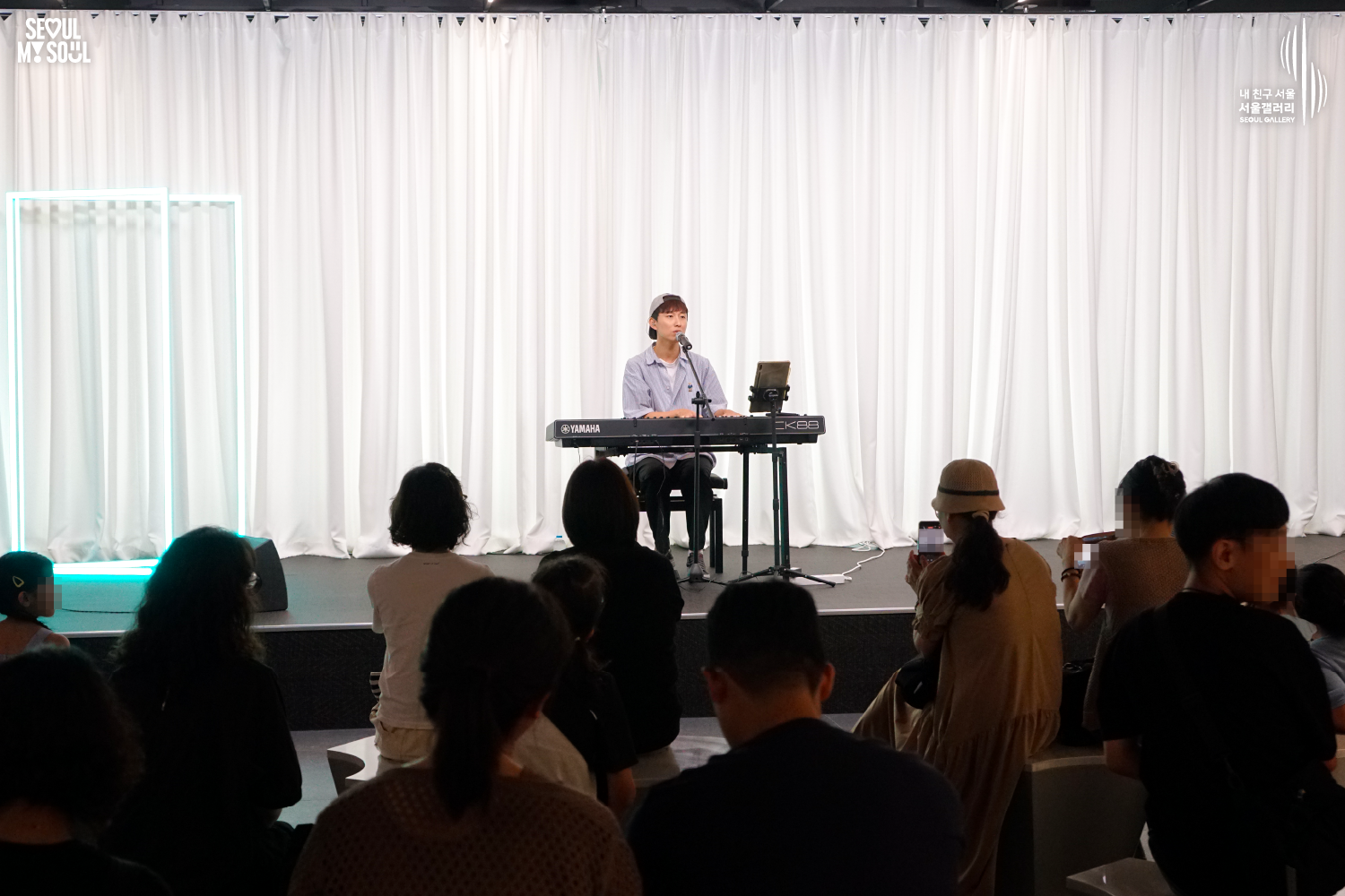 SEOUL MY SOUL 로고와 내 친구 서울 서울갤러리 SEOUL GALLERY 로고가 표기된 서울갤러리 공연장 사진으로, 흰색 커튼과 청록빛 사각 네온 조명이 보이는 무대 위에서 모자를 뒤로 쓴 짧은 머리의 남성 연주자가 의자에 앉아 키보드를 연주하며 마이크 앞에서 노래를 부르고 있으며, 무대 위에는 마이크 스탠드와 태블릿 거치대, 모니터 스피커가 놓여 있고 객석에는 관람객들이 공연을 관람하며 일부는 휴대전화로 무대를 촬영하고 있는 모습이 담긴 이미지입니다.