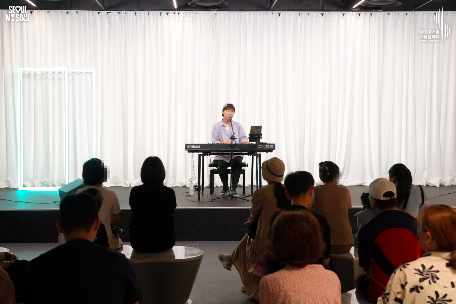 SEOUL MY SOUL 로고와 내 친구 서울 서울갤러리 SEOUL GALLERY 로고가 표기된 서울갤러리 공연장 사진으로, 흰색 커튼과 청록빛 사각 네온 조명이 보이는 무대 위에서 모자를 뒤로 쓴 짧은 머리의 남성 연주자가 의자에 앉아 키보드를 연주하며 마이크 앞에서 노래를 부르고 있으며, 무대 위에는 마이크 스탠드와 태블릿 거치대, 모니터 스피커가 놓여 있고 전경에는 다양한 연령대의 관람객들이 앉아 공연을 관람하고 있는 모습이 담긴 이미지입니다.