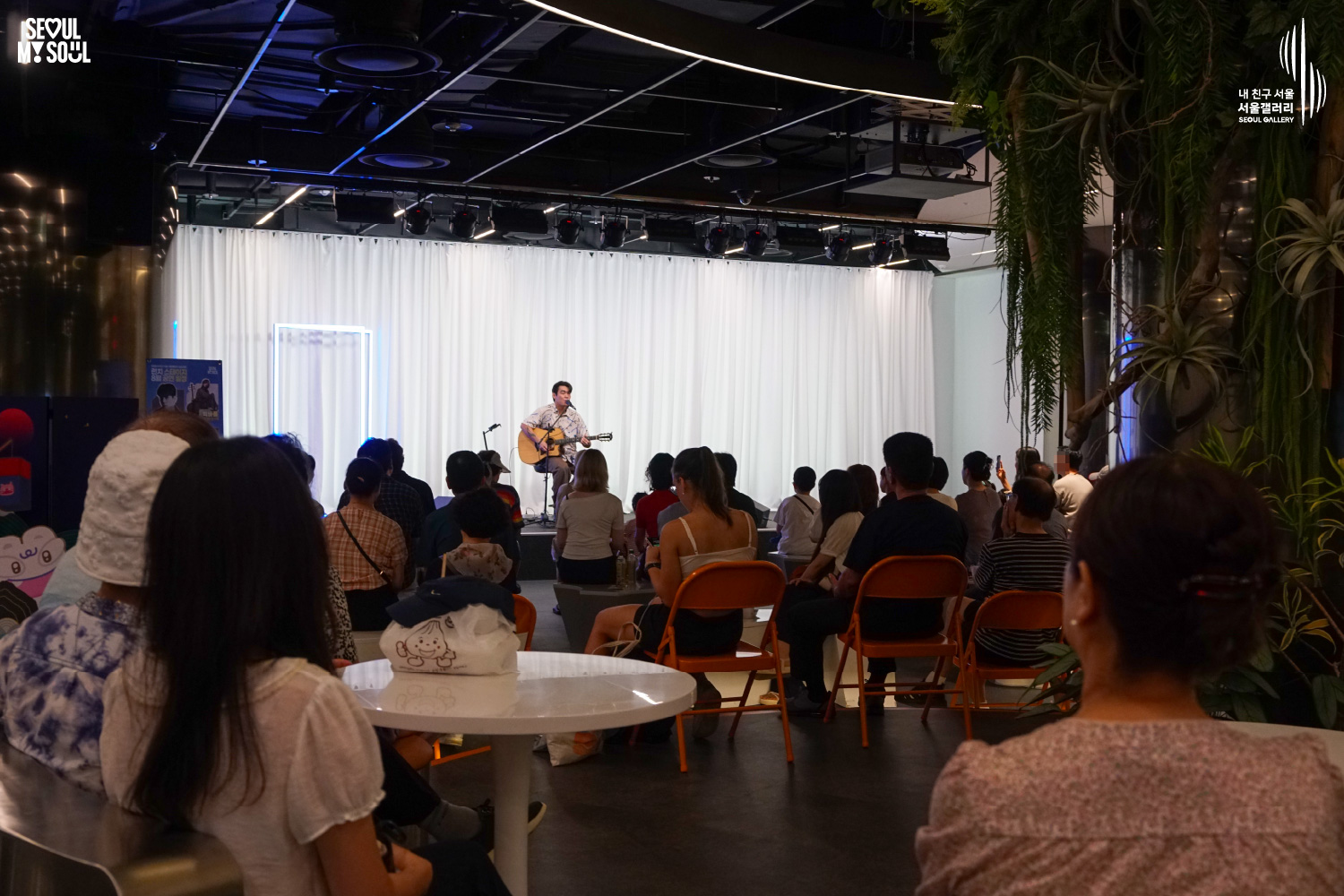 SEOUL MY SOUL 로고와 내 친구 서울 서울갤러리 SEOUL GALLERY 로고가 표기된 서울갤러리 지하 1층 공연장의 공연 현장 사진으로, 천장에 무대 조명이 설치되고 흰색 커튼과 푸른빛 사각 네온 조명이 보이는 무대 위에서 짧은 머리의 남성 싱어송라이터가 의자에 앉아 어쿠스틱 기타를 연주하며 마이크 앞에서 노래를 부르고 있으며, 객석에는 다양한 연령대의 관람객들이 앉아 공연을 관람하고 있는 모습이 담긴 이미지입니다.