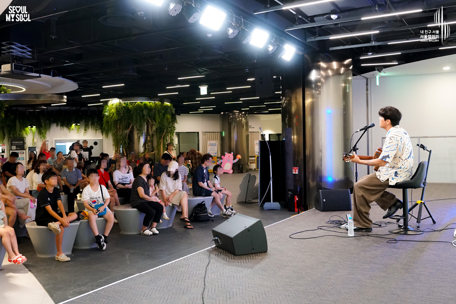 SEOUL MY SOUL 로고와 내 친구 서울 서울갤러리 SEOUL GALLERY 로고가 표기된 서울갤러리 지하 1층 공연장의 공연 현장 사진으로, 천장에 무대 조명이 설치된 실내 공간에서 짧은 머리의 남성 싱어송라이터가 의자에 앉아 어쿠스틱 기타를 연주하며 마이크 앞에서 노래를 부르고 있으며, 무대 앞에는 모니터 스피커가 놓여 있고 객석에는 어린이를 포함한 다양한 연령대의 관람객들이 앉아 공연을 관람하고 있는 모습이 담긴 이미지입니다.