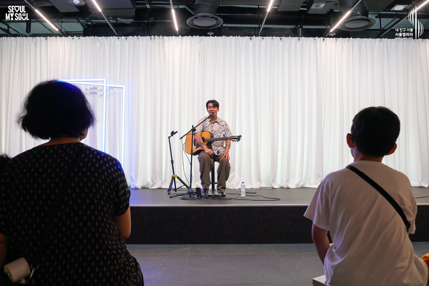 SEOUL MY SOUL 로고와 내 친구 서울 서울갤러리 SEOUL GALLERY 로고가 표기된 서울갤러리 공연장 사진으로, 천장에 조명이 설치되고 흰색 커튼과 푸른빛 사각 네온 조명이 보이는 무대 위에서 짧은 머리의 남성 싱어송라이터가 의자에 앉아 어쿠스틱 기타를 든 채 마이크 앞에서 관객을 바라보며 이야기하고 있으며, 전경에는 무대를 바라보는 관람객 두 명의 뒷모습이 보이는 모습이 담긴 이미지입니다.