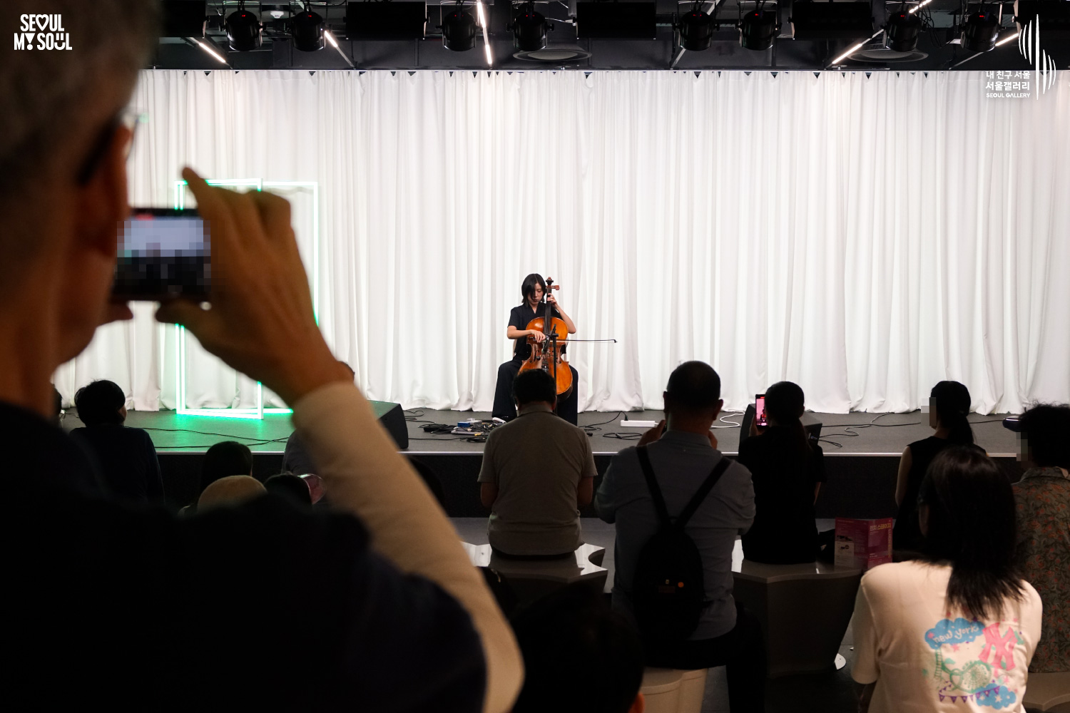 SEOUL MY SOUL 로고와 내 친구 서울 서울갤러리 SEOUL GALLERY 로고가 표기된 서울갤러리 공연장 사진으로, 천장에 무대 조명이 설치되고 흰색 커튼과 초록빛 사각 네온 조명이 보이는 무대 위에서 단발머리의 여성 연주자가 의자에 앉아 첼로를 활로 연주하고 있으며, 객석에는 다양한 연령대의 관람객들이 앉아 공연을 관람하고 일부 관람객은 휴대전화로 무대를 촬영하고 있는 모습이 담긴 이미지입니다.