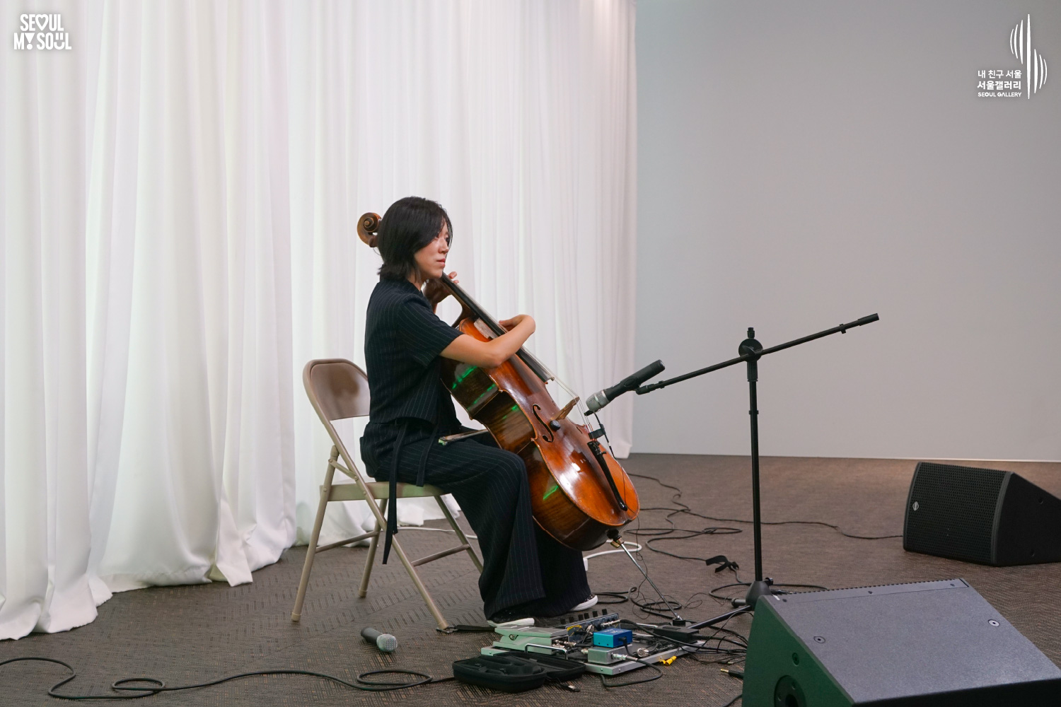 SEOUL MY SOUL 로고와 내 친구 서울 서울갤러리 SEOUL GALLERY 로고가 표기된 서울갤러리 공연장 무대 사진으로, 은은한 조명이 비추는 흰색 커튼 배경의 무대 위에서 단발머리의 여성 연주자가 의자에 앉아 첼로를 연주하고 있으며, 무대 위에는 마이크 스탠드와 페달 이펙터, 모니터 스피커가 놓여 있고 바닥에는 케이블이 연결되어 있는 모습이 담긴 이미지입니다.