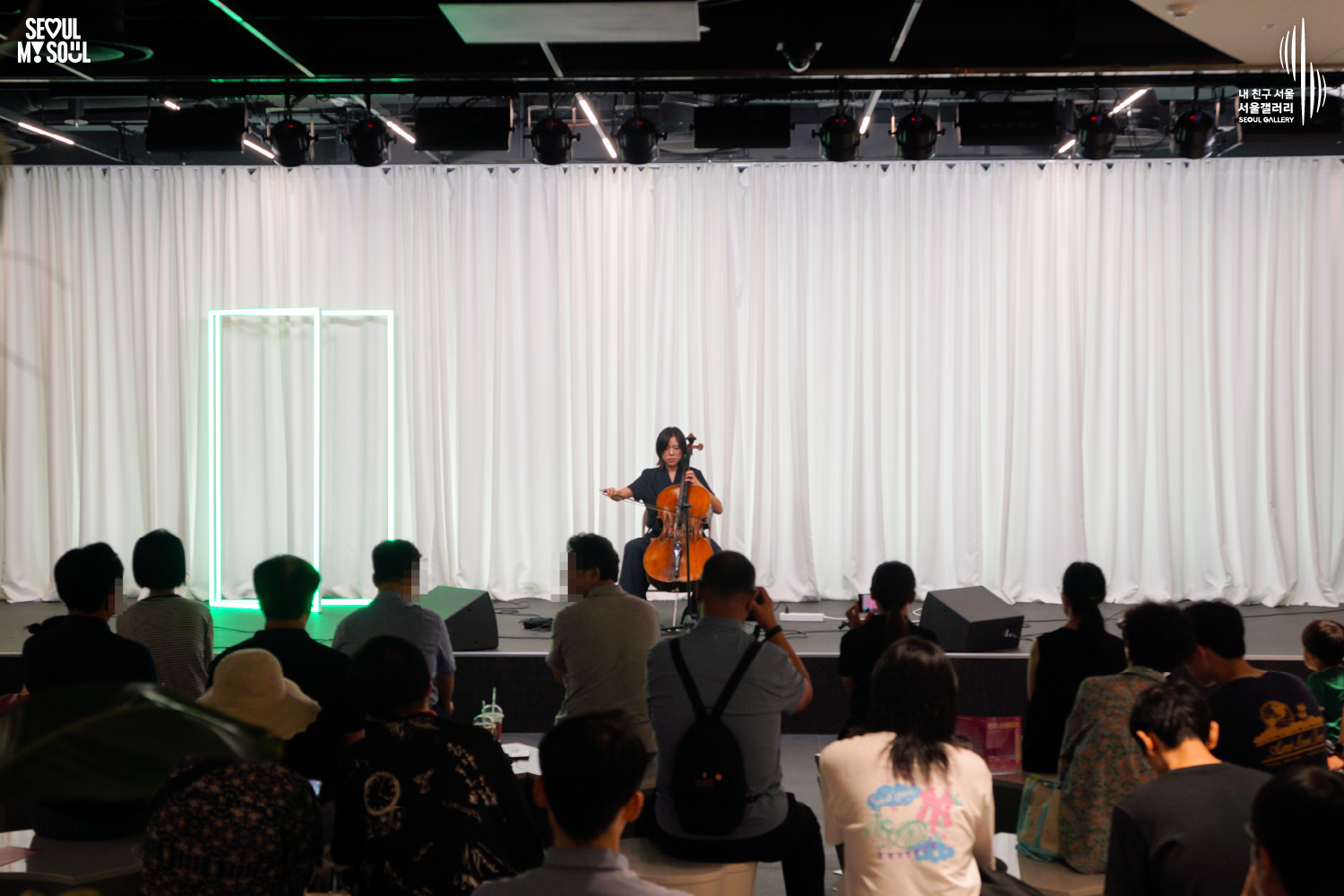 SEOUL MY SOUL 로고와 내 친구 서울 서울갤러리 SEOUL GALLERY 로고가 표기된 서울갤러리 지하 1층 공연장의 공연 현장 사진으로, 천장에 무대 조명이 설치되고 흰색 커튼과 초록빛 사각 네온 조명이 보이는 무대 위에서 단발머리의 여성 연주자가 의자에 앉아 첼로를 연주하고 있으며, 객석에는 다양한 연령대의 관람객들이 앉아 공연을 관람하고 일부 관람객은 휴대전화로 무대를 촬영하고 있는 모습이 담긴 이미지입니다.