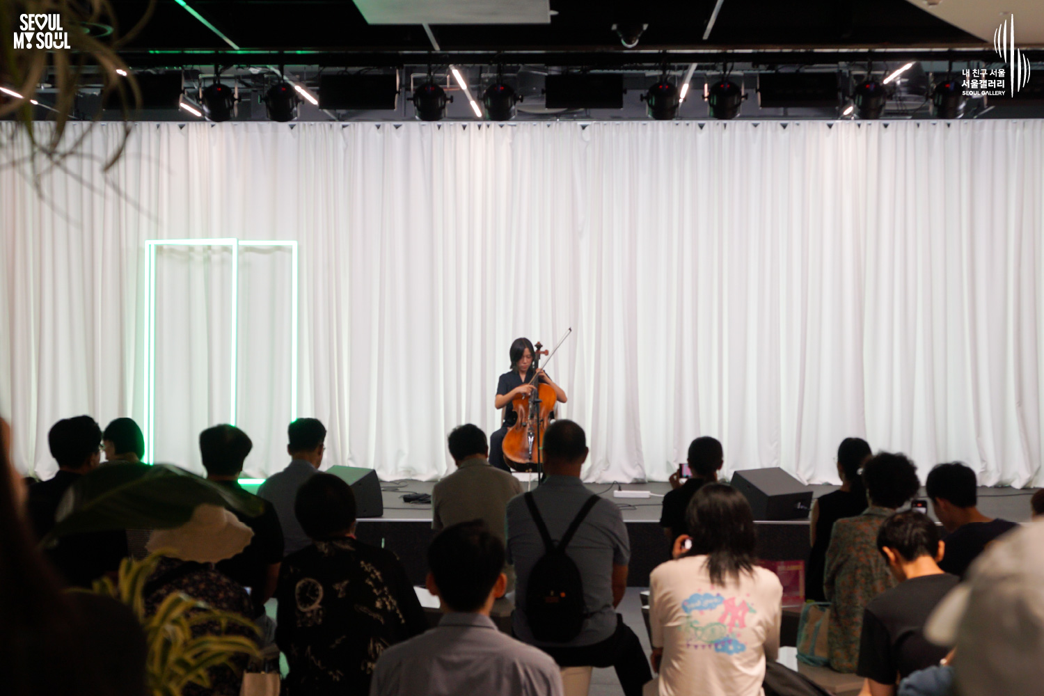SEOUL MY SOUL 로고와 내 친구 서울 서울갤러리 SEOUL GALLERY 로고가 표기된 서울갤러리 지하 1층 공연장의 공연 현장 사진으로, 천장에 무대 조명이 설치되고 흰색 커튼과 초록빛 사각 네온 조명이 보이는 무대 위에서 단발머리의 여성 연주자가 의자에 앉아 첼로를 활로 연주하고 있으며, 객석에는 다양한 연령대의 관람객들이 앉아 공연을 관람하고 일부 관람객은 휴대전화로 무대를 촬영하고 있는 모습이 담긴 이미지입니다.